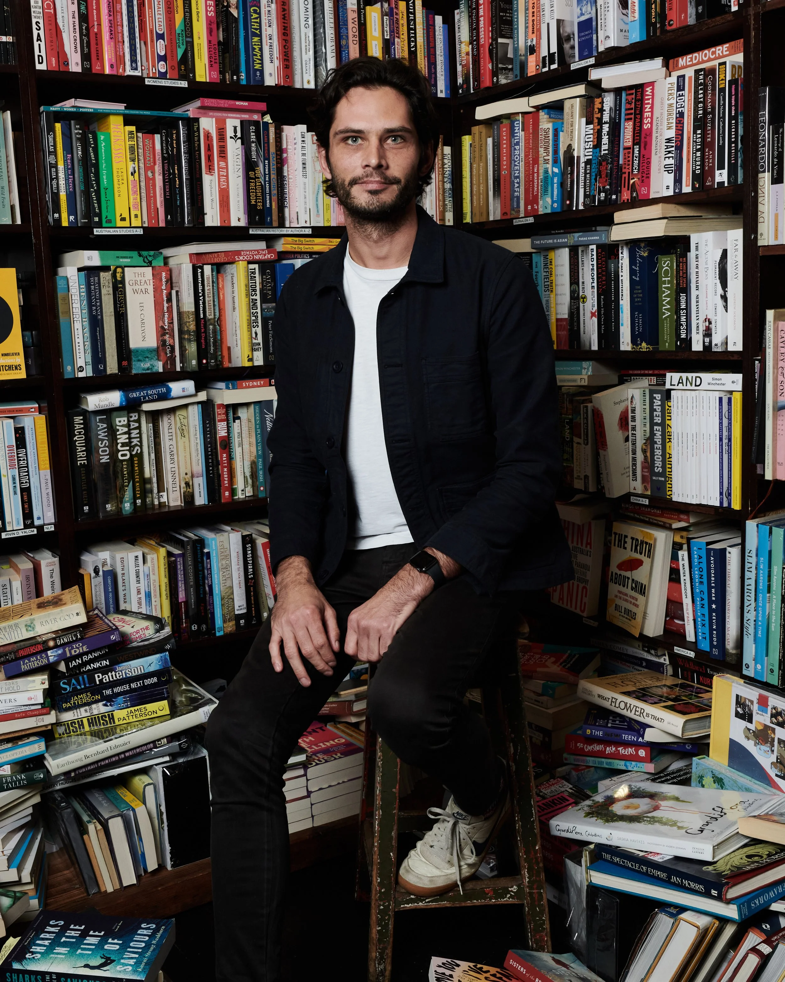 A man with dark hair and a beard sits in front of bookshelves filled with various books, wearing a black jacket and white shirt, with one leg on a step stool and the other on the ground in a bookstore or library.