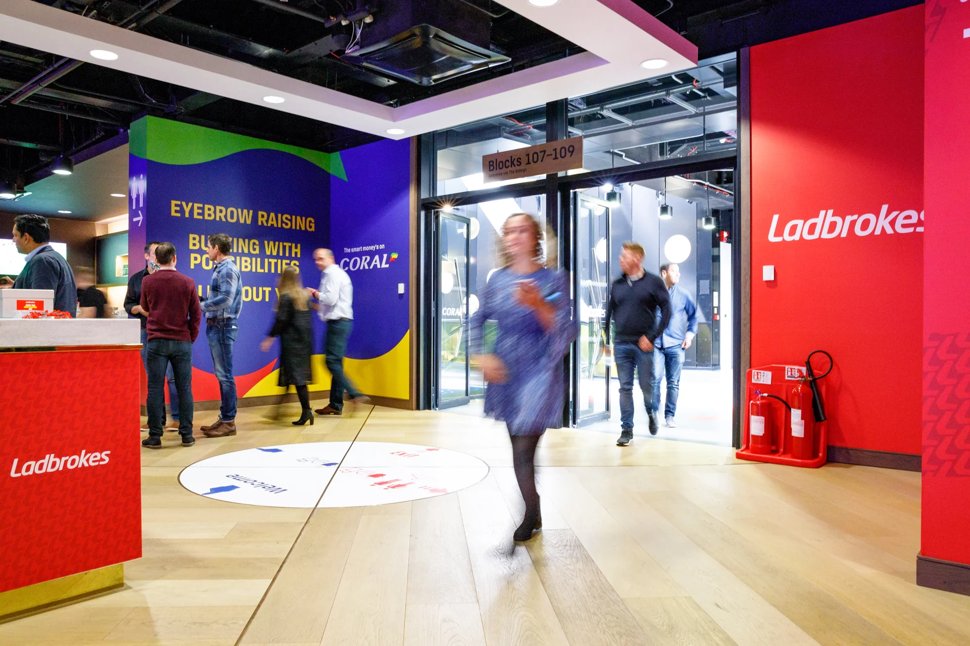 People walking into a building with blue, yellow, and red walls, and a sign indicating blocks 107-109. The floor has a circular game or seating area with the word 'welcome' and arrows. There are fire extinguishers and ceiling lights visible.