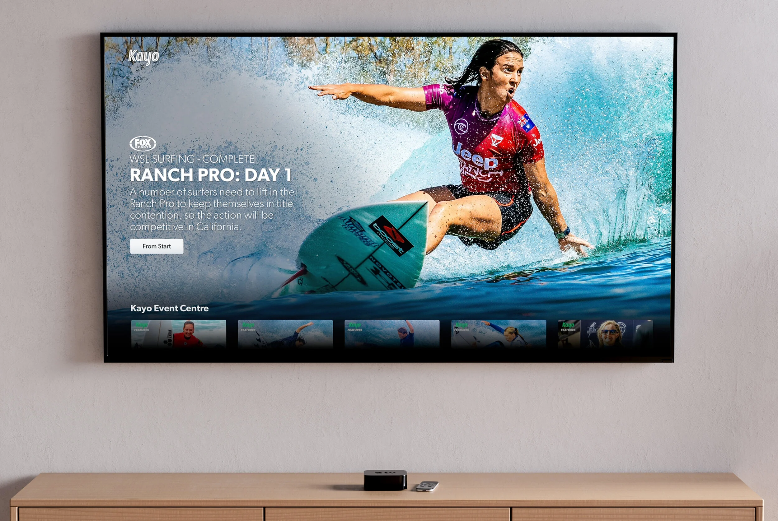 Television screen displaying a surfing competition with a female surfer in a purple and red wetsuit riding a wave, with text about the event titled 'Ranch Pro: Day 1' hosted at Kayo Event Centre.