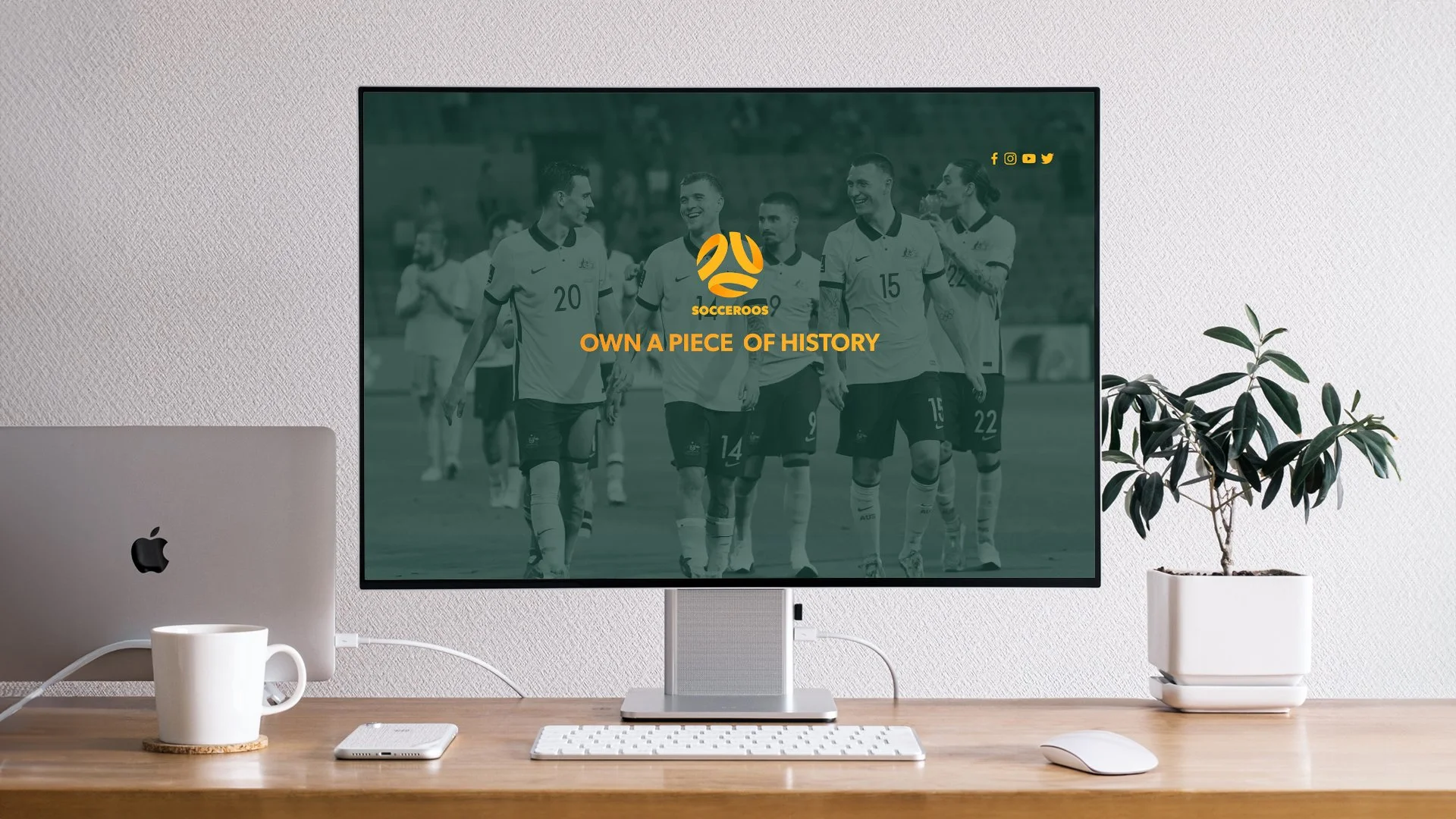 Computer monitor displaying a soccer team celebrating, with the text 'OWN A PIECE OF HISTORY' and the logo 'SOCCEROOS'.