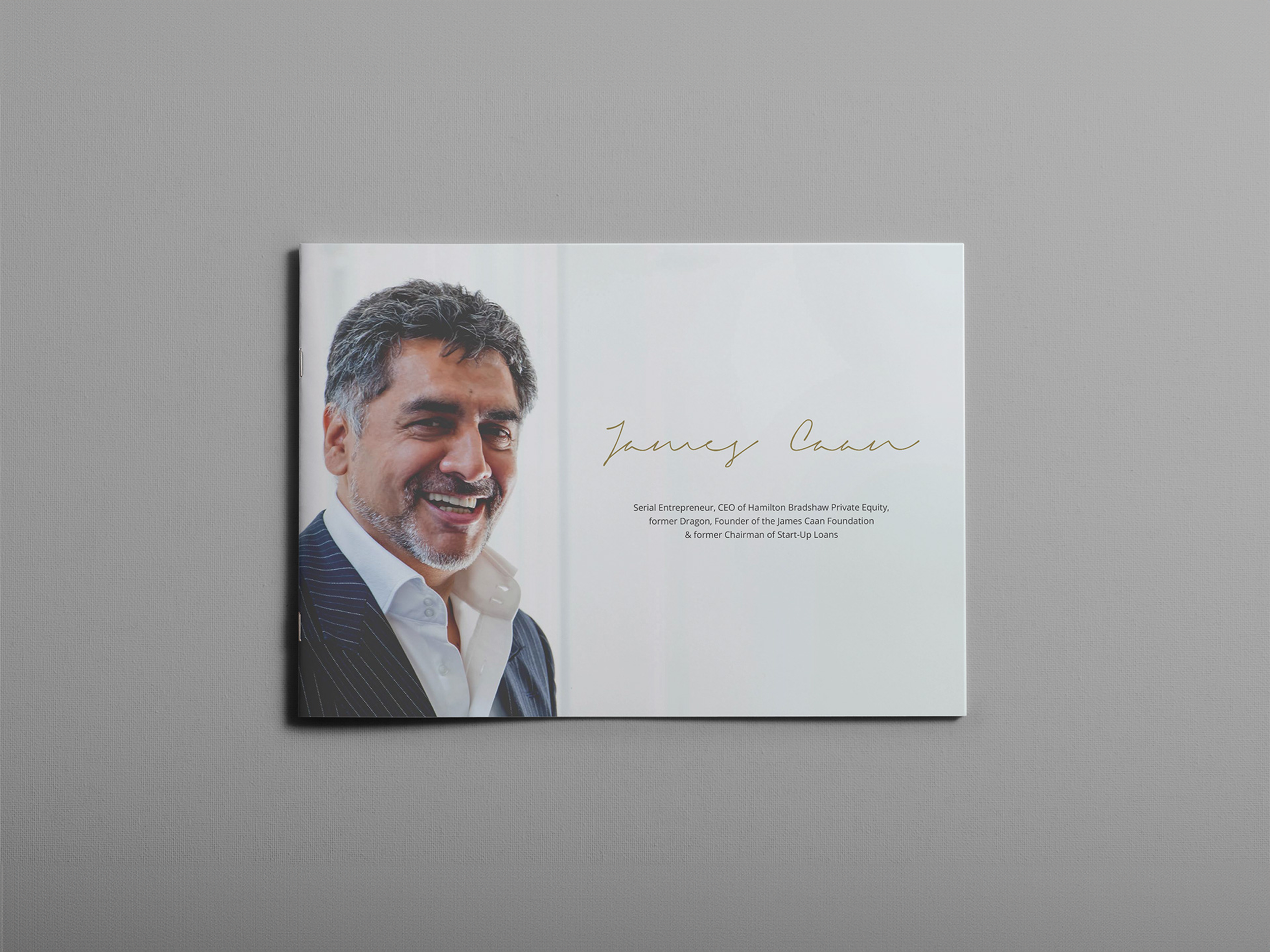 A magazine or brochure featuring a smiling man with gray hair and a beard, dressed in a dark pinstripe suit and white shirt, with gold-colored cursive text on the page.