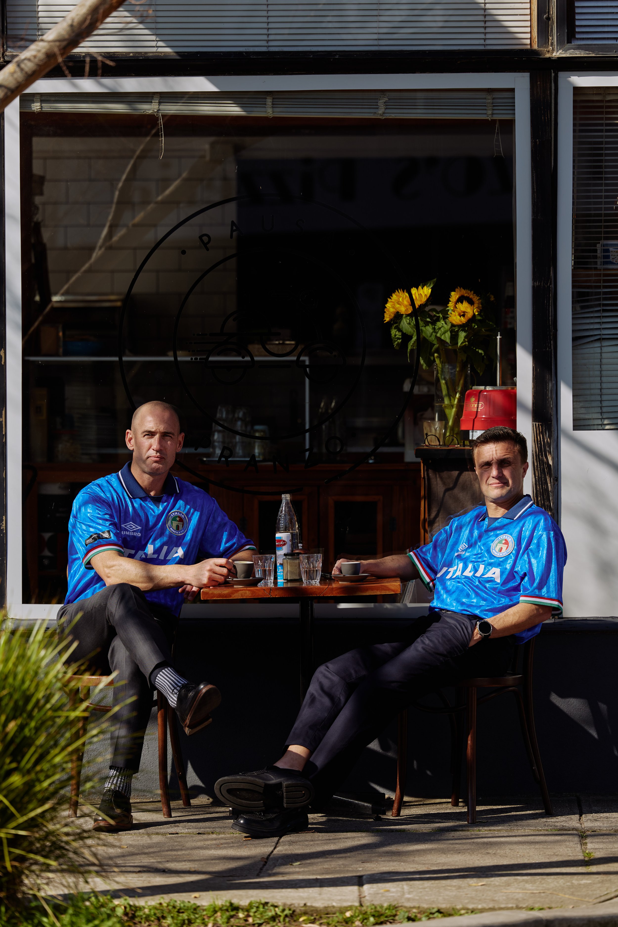 Two men wearing blue Umbro Italia football shirts seated at a cafe table
