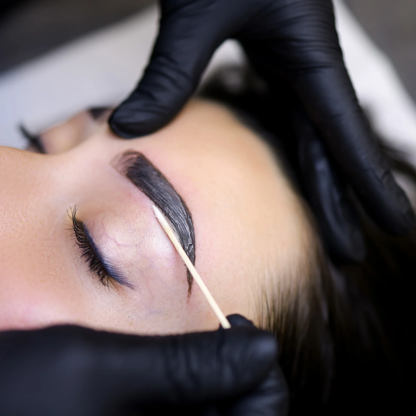 A person receiving an eyebrow tattoo or microblading procedure, with black gloves manipulating the eyebrow area and a cotton swab applied to the skin.