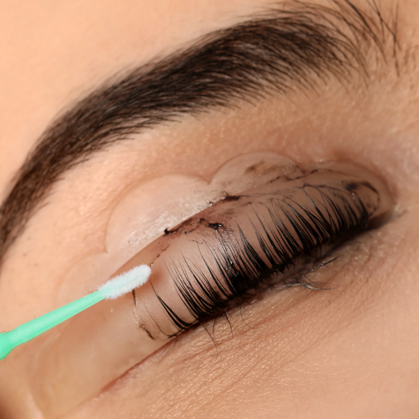 Close-up of a person with long, curled eyelashes and neatly groomed eyebrow, as a cotton swab is being used to apply adhesive to the eyelash extension.