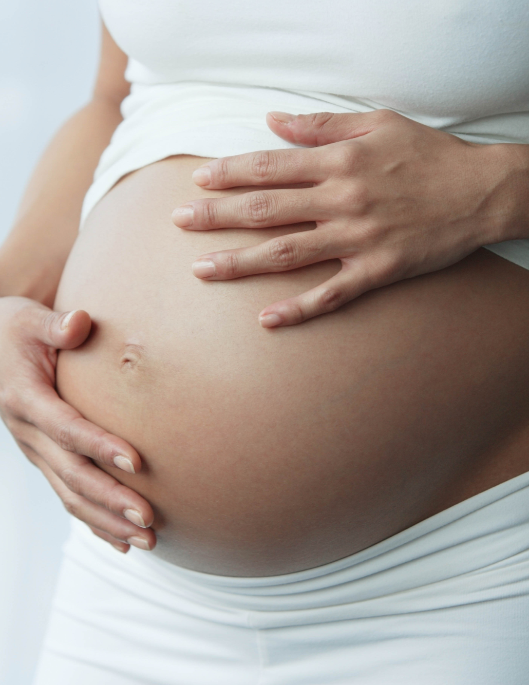 Close-up of pregnant woman's belly with hands gently resting on it, wearing a white top.