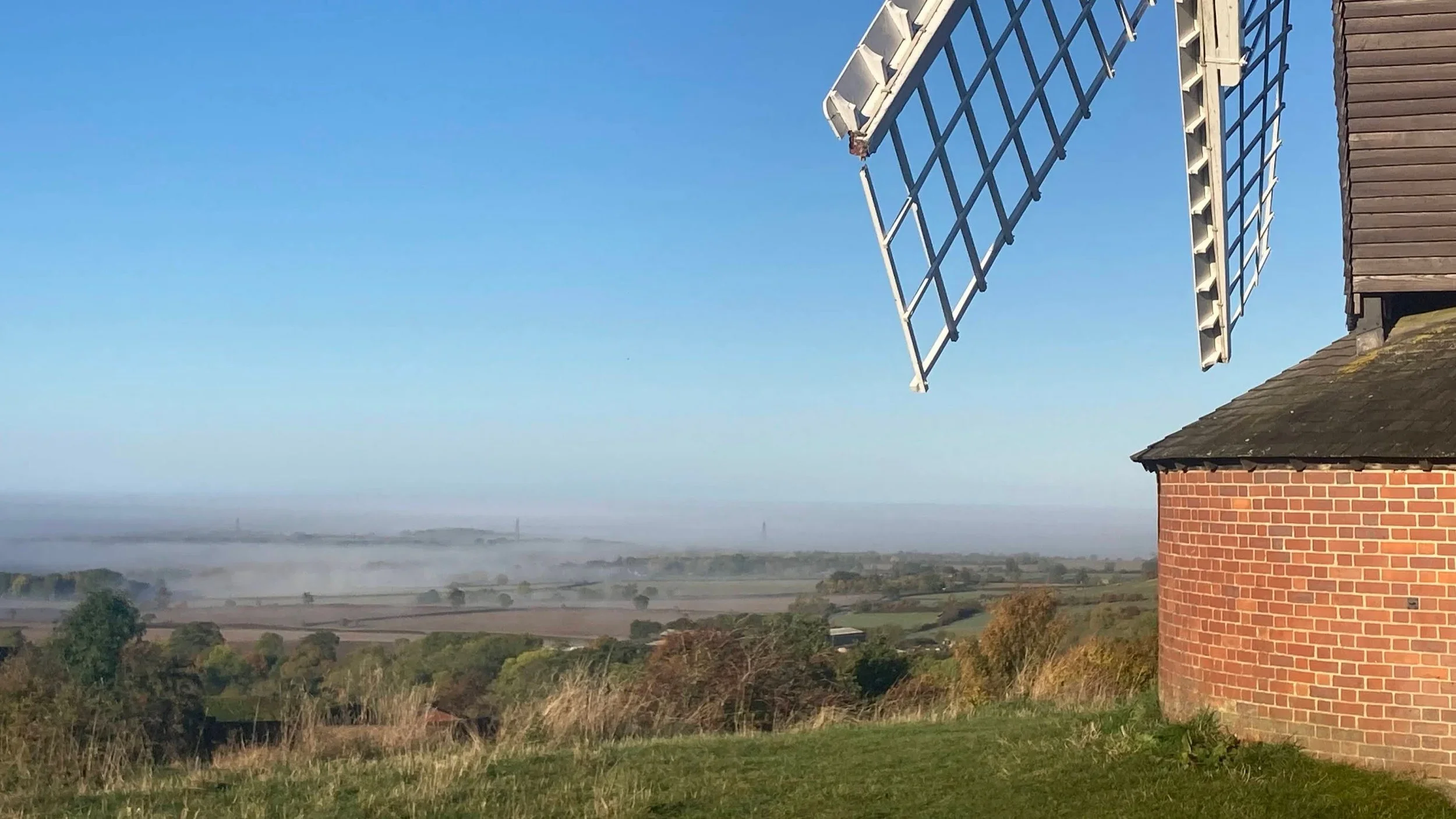 Photo of distance mist-shrouded fields and woods with part of Brill Windmill in the foreground.