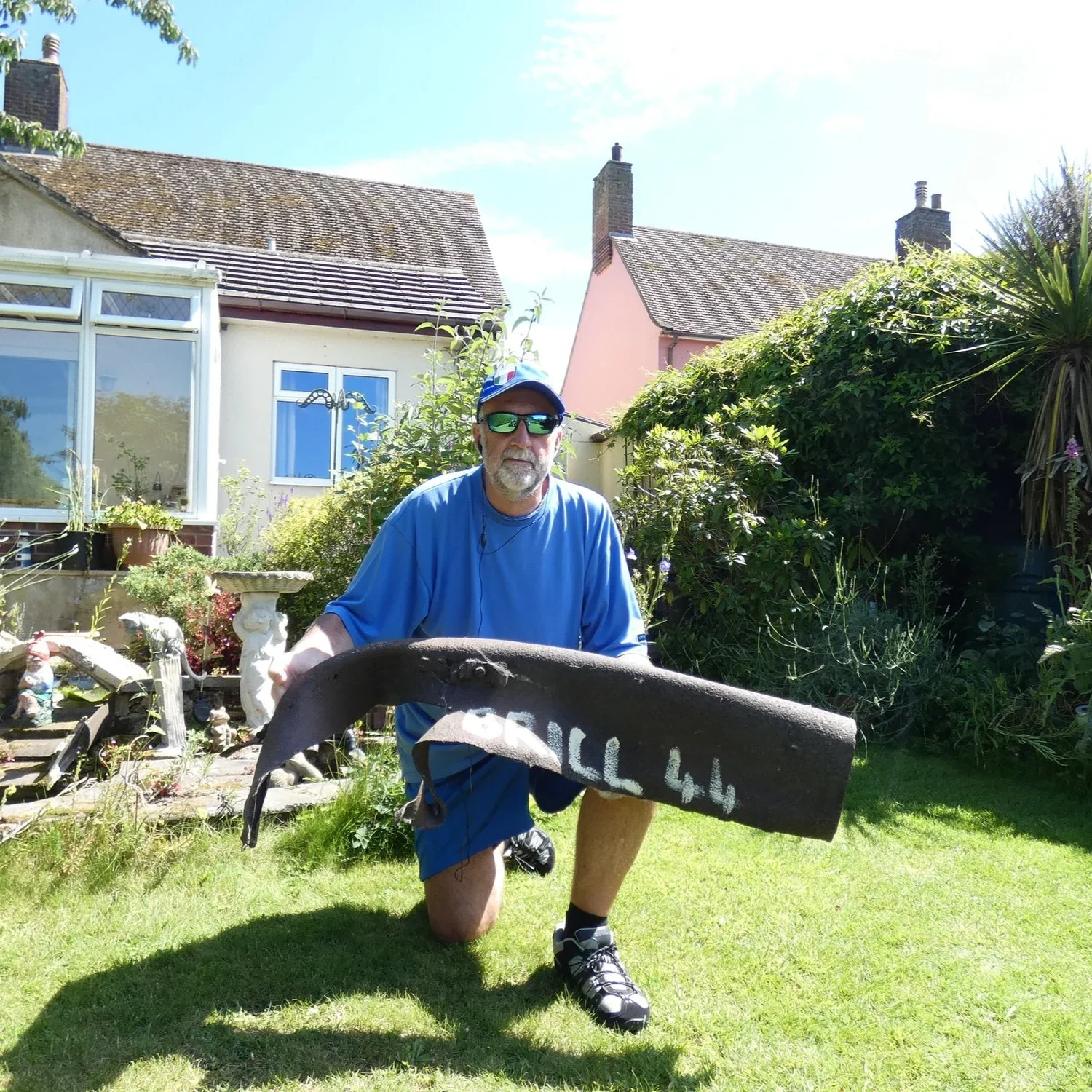 Photo of a man kneeling in a sunny gardens holding a large piece of grey metal.