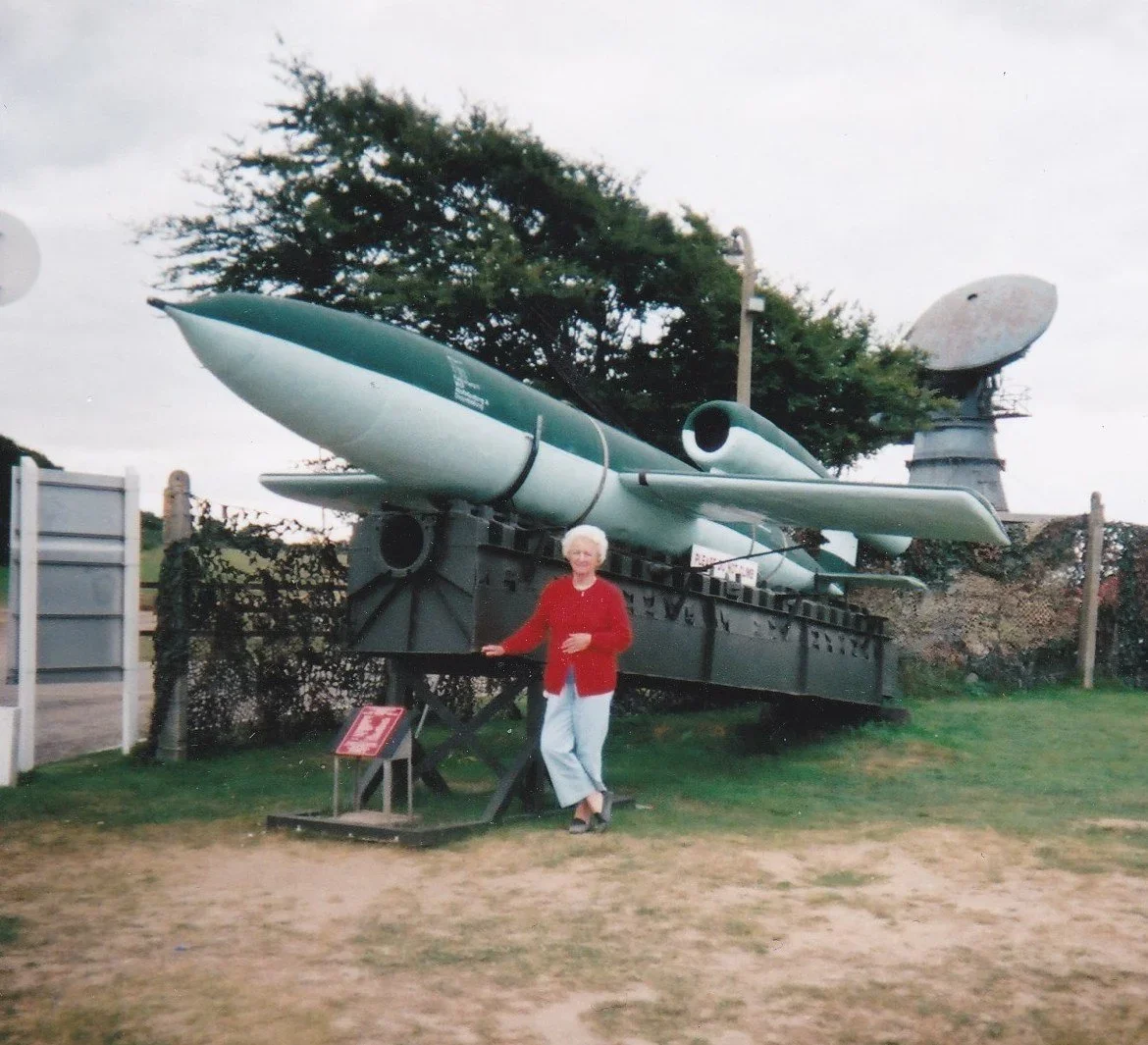 Photo of a white-haired woman is a red cardigan posing next to a large green missile.