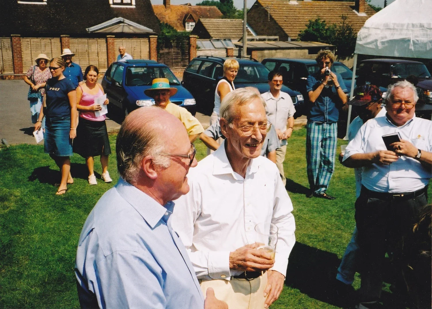 Photo of two older men standing chatting in the open air with other people looking on.