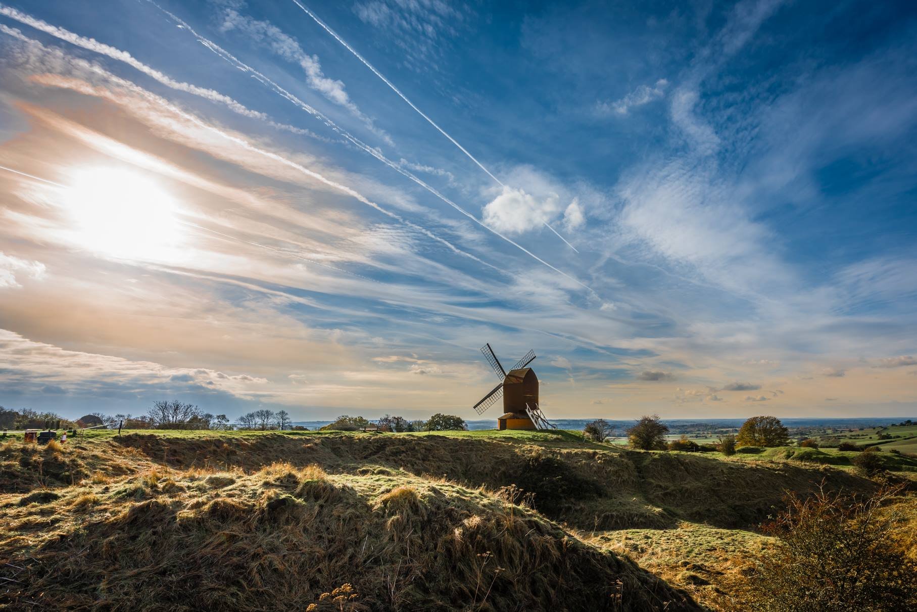 Photo of Brill Windmill in the distance with grass in the foreground and a dramatic sky of clouds and contrails.