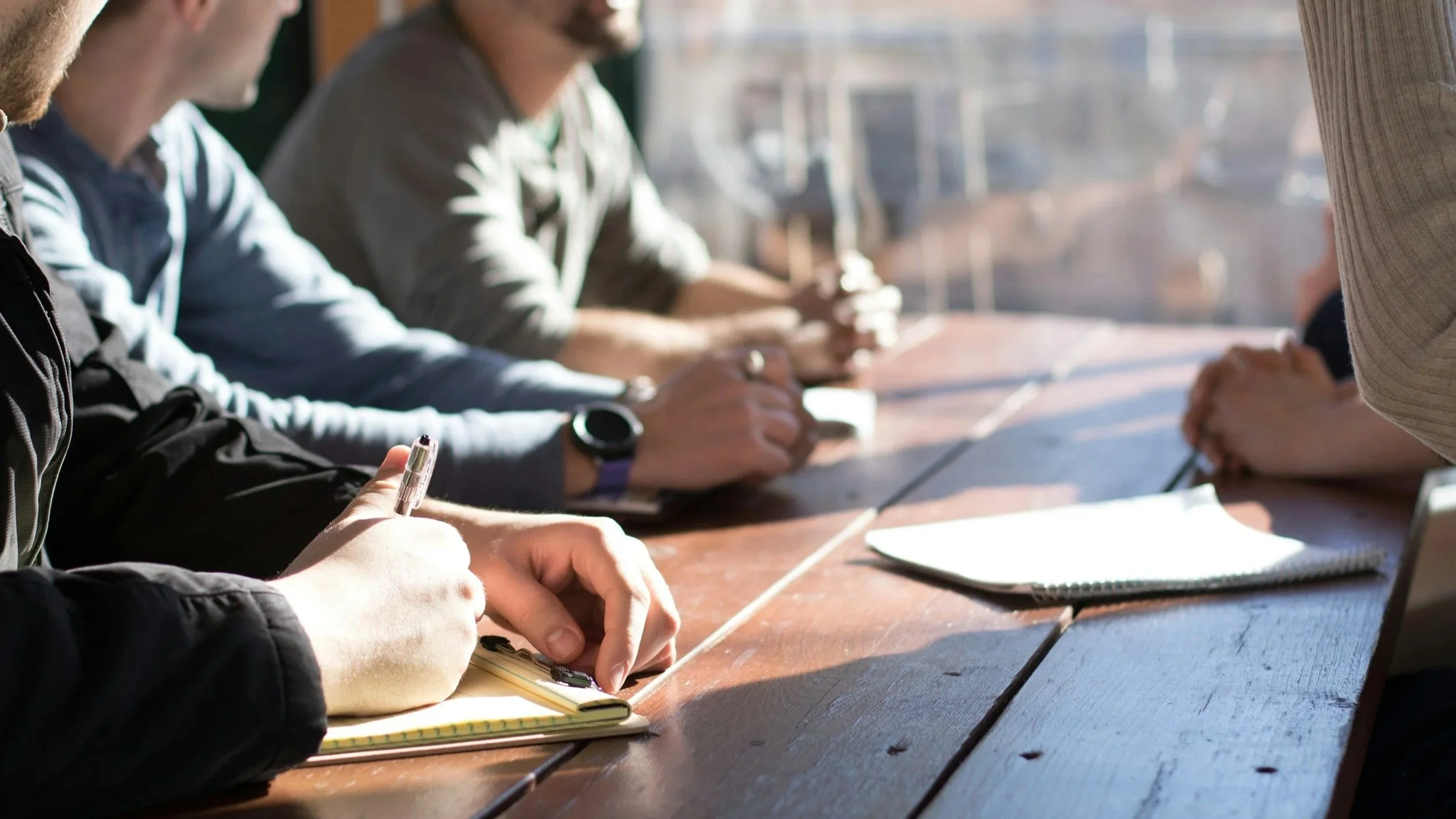Stock image of four sets of hands and notepads resting on a table.