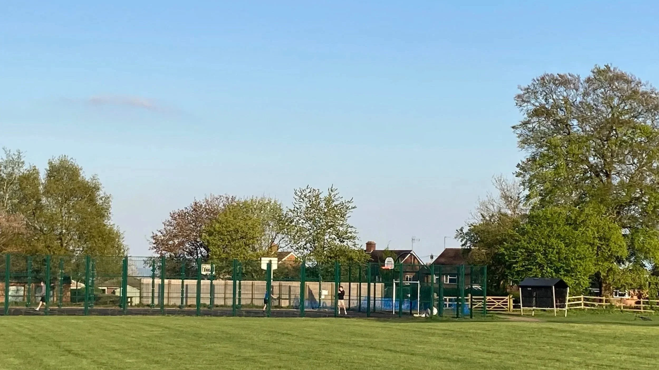 Photo of fenced play area with trees and houses behind under blue sky.