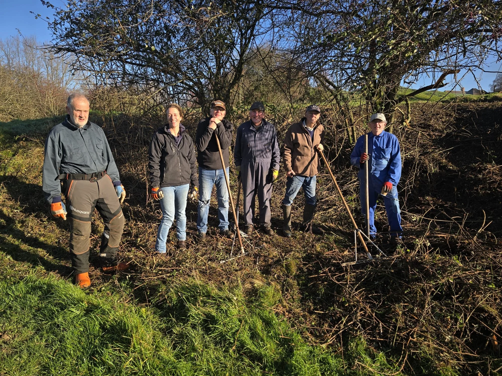 Photo of 5 men and one woman in working clothes holding tools surrounded by rough grass and winter trees.