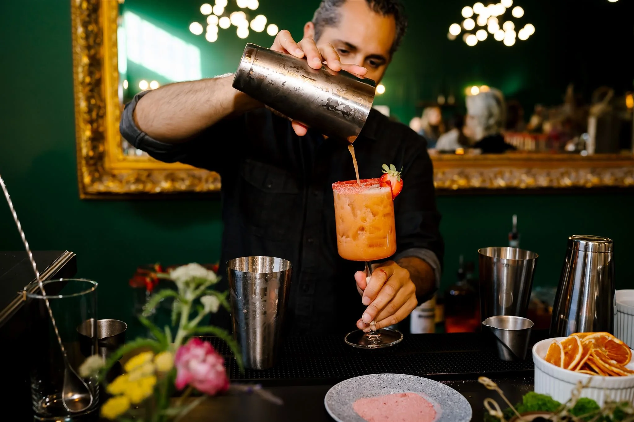 Bartender pouring cocktail from a shaker into a glass with a strawberry garnish, surrounded by cocktail tools and ingredients.