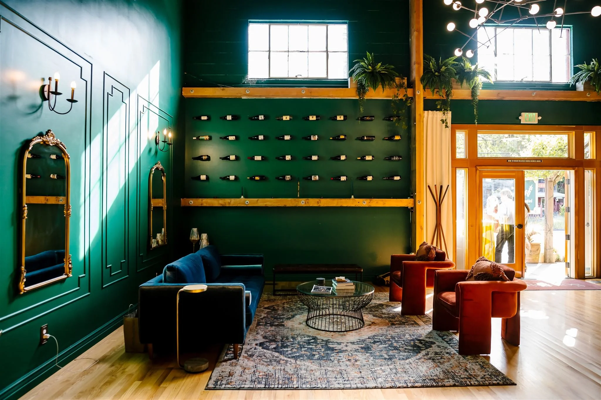 Stylish living room with dark green walls, blue sofa, red chairs, decorative mirrors, wall-mounted wine bottles, and modern chandelier.