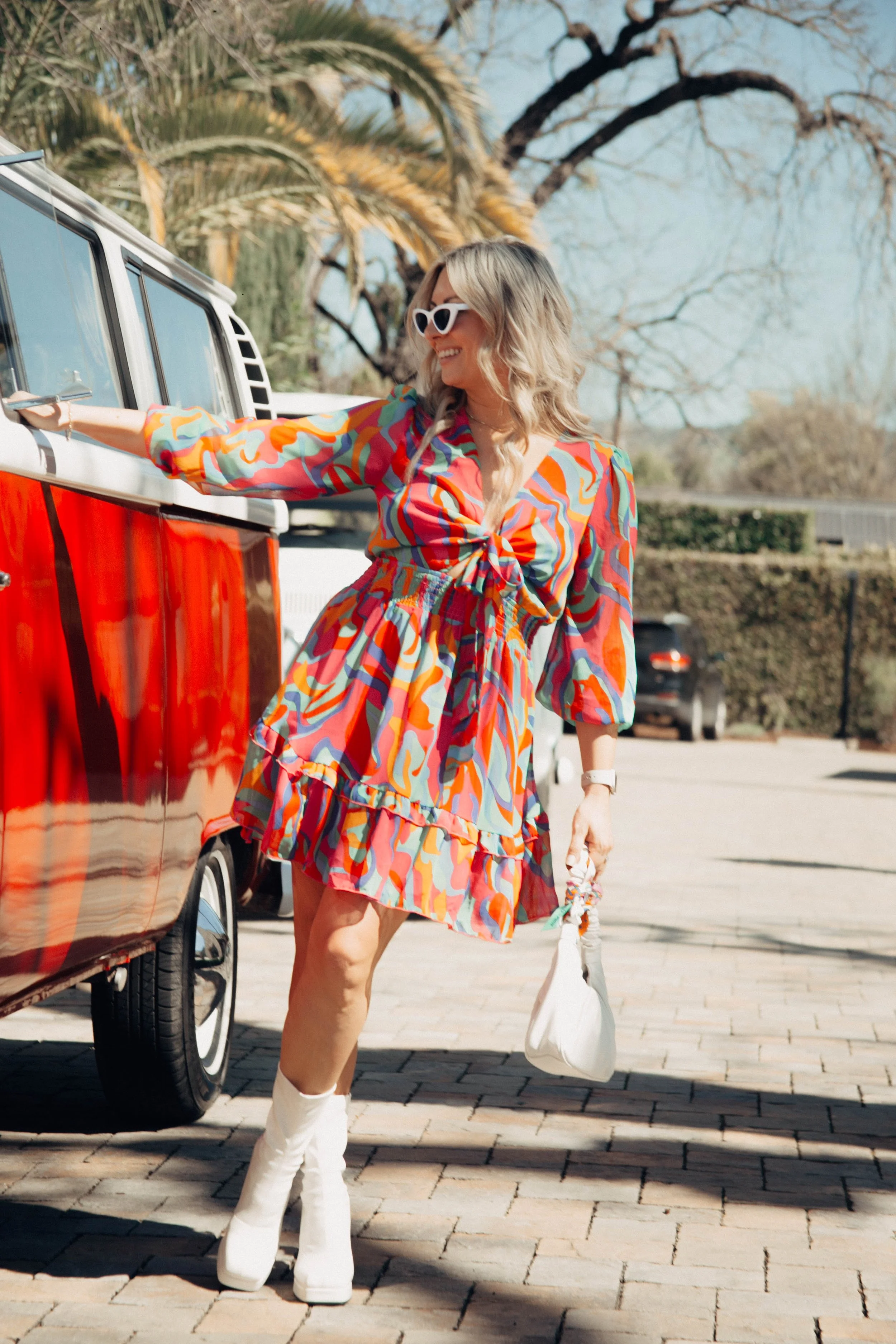Woman in colorful dress with white boots, wearing white sunglasses, standing next to a red and white vehicle, smiling and holding a small bag.