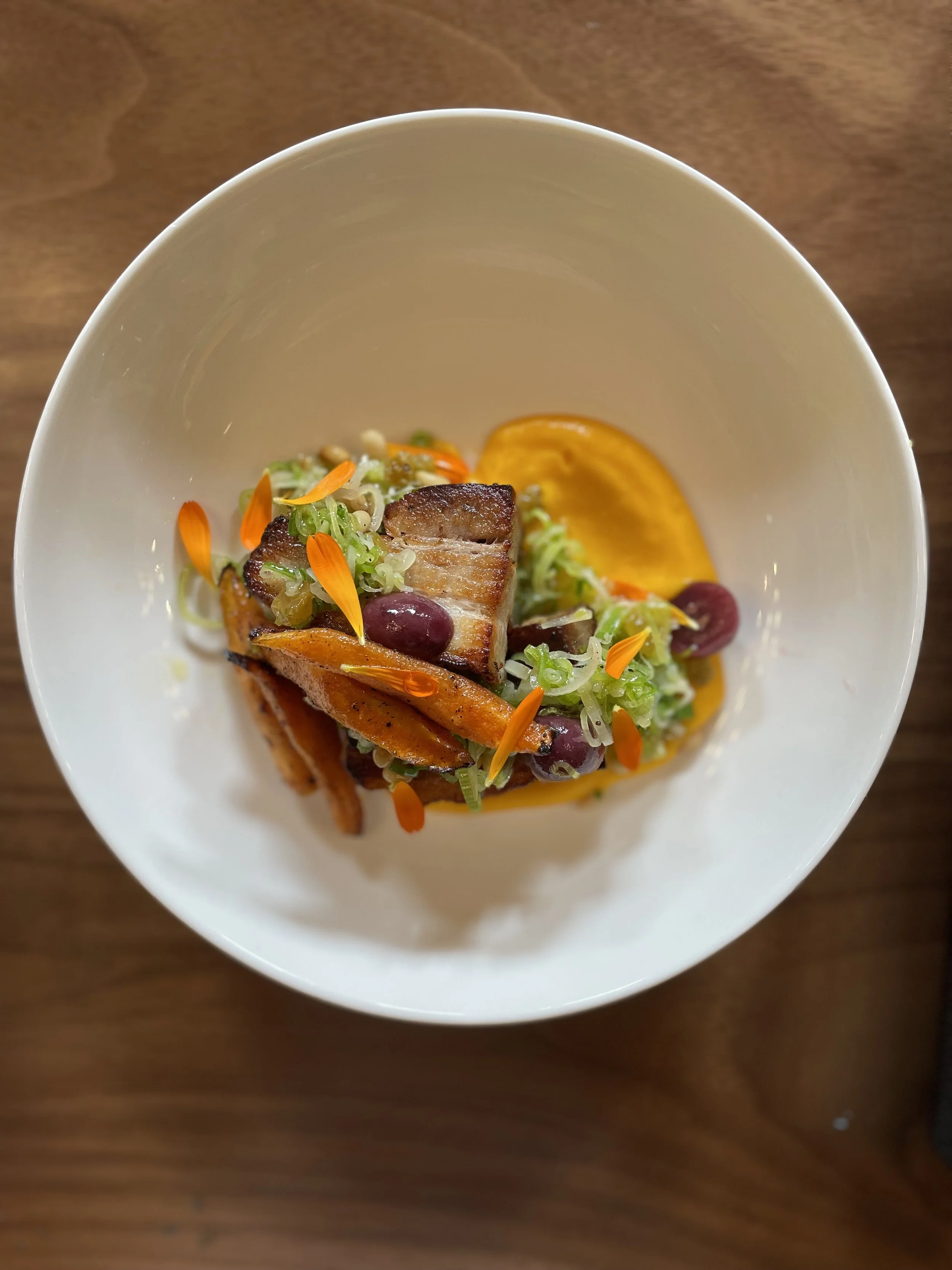 Gourmet dish with grilled meat, vegetables, and sauce in a white bowl on a wooden table.