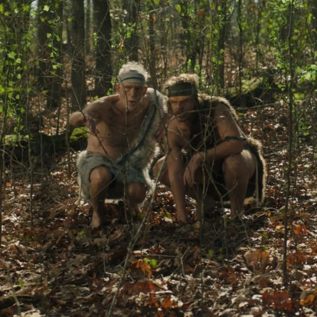 Two men in primitive costumes crouching in the woods during the “Human Food Bar” commercial.