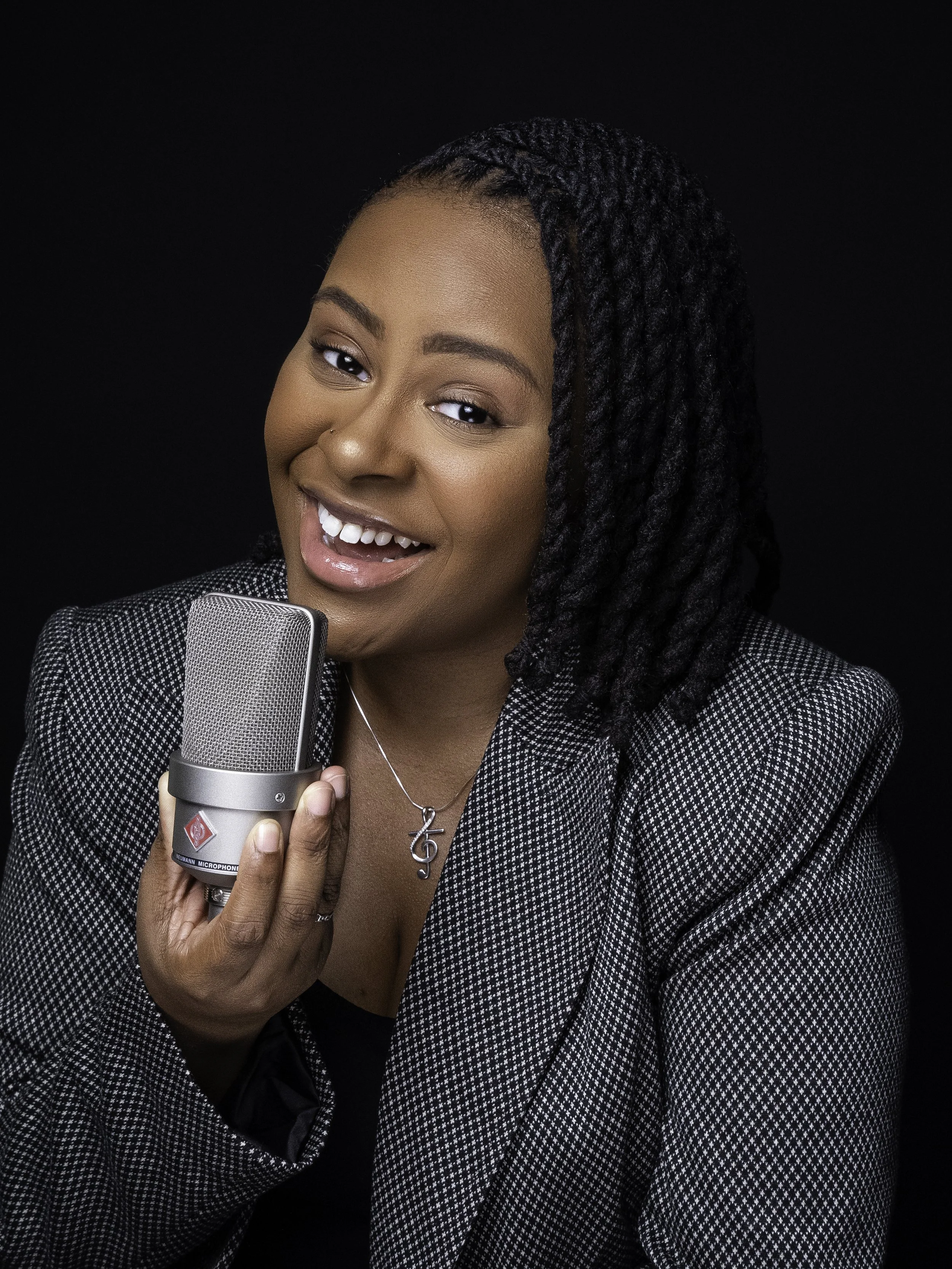 A woman with dark skin and braided hair holding a microphone, dressed in a checkered blazer, smiling at the camera against a black background.