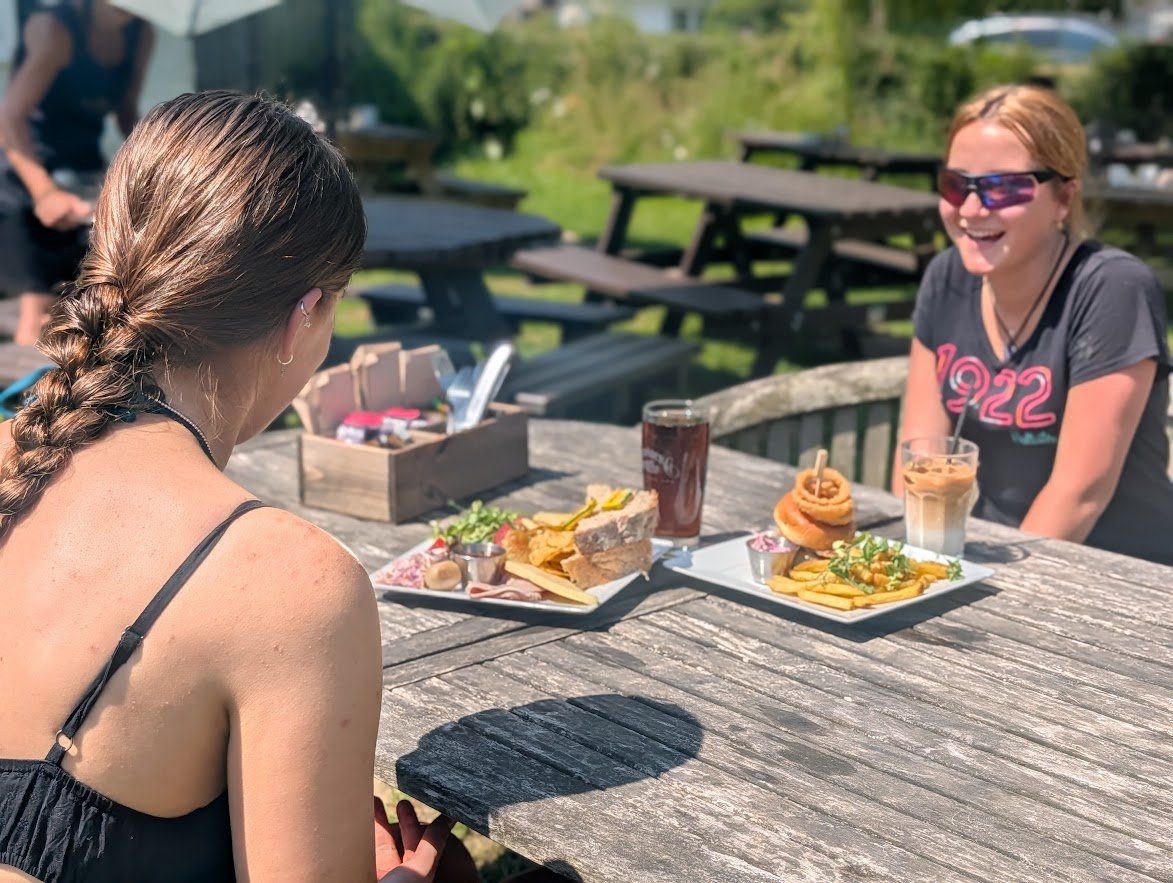 Two women sitting at a wooden picnic table outdoors on a sunny day, enjoying lunch with plates of food, drinks, and condiments.