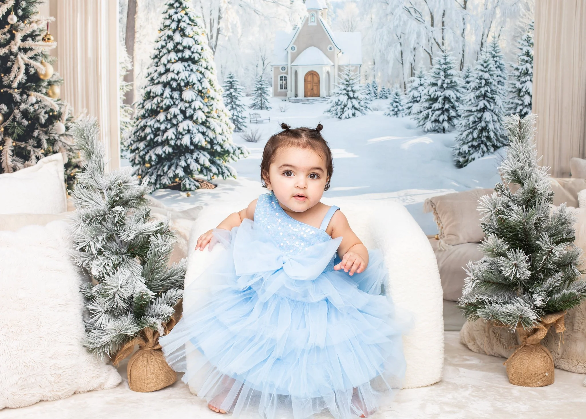 A young girl dressed as a princess in a blue gown sitting indoors in front of a winter scene with snow-covered trees, a church, and Christmas trees decorated with ornaments.