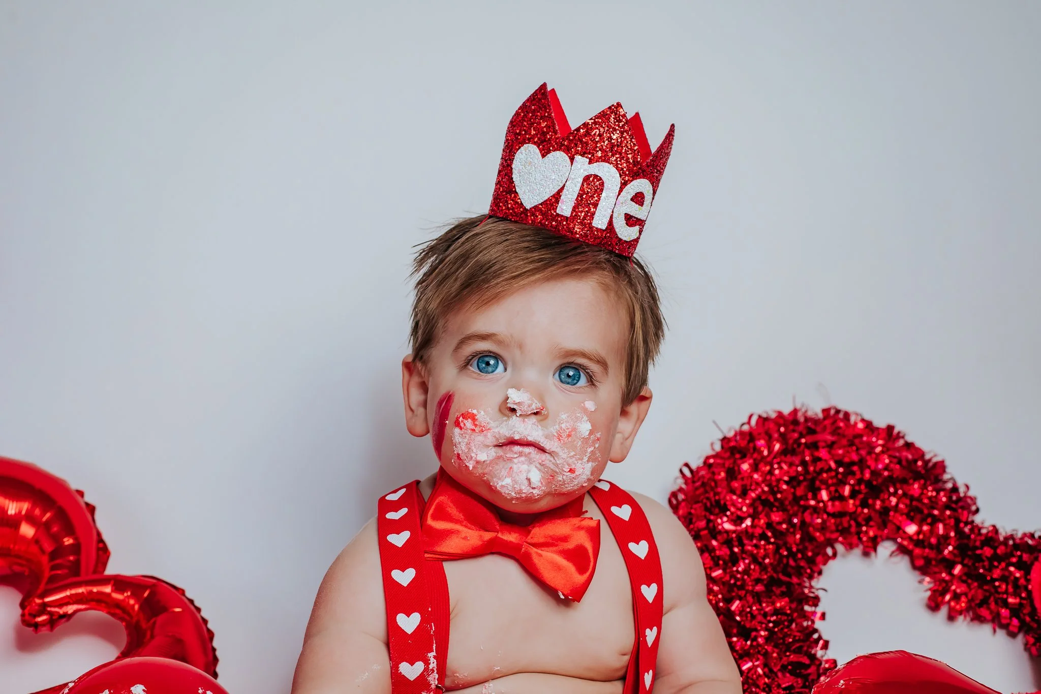 A young child with blue eyes and light brown hair, wearing a red and white heart-patterned suspenders, red bow tie, and a red heart-shaped headband with the word 'me' on it, has their face covered in white and red cake or frosting and is surrounded b