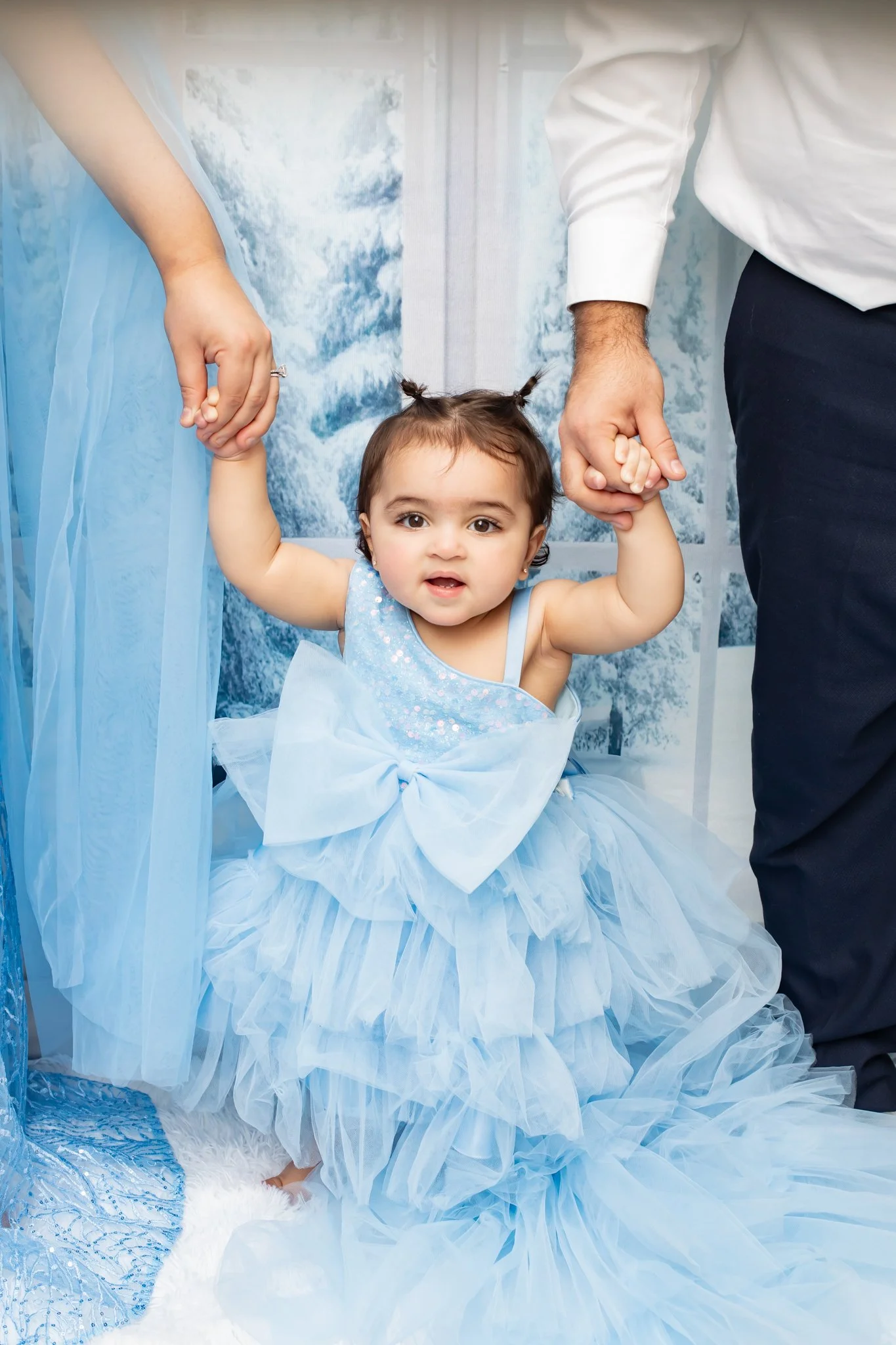 A young girl dressed in a blue princess costume with tulle layers is sitting on a white fluffy rug, holding hands with two adults, one in a white shirt and the other in a blue dress. The girl has dark hair styled in two small pigtails and is looking 