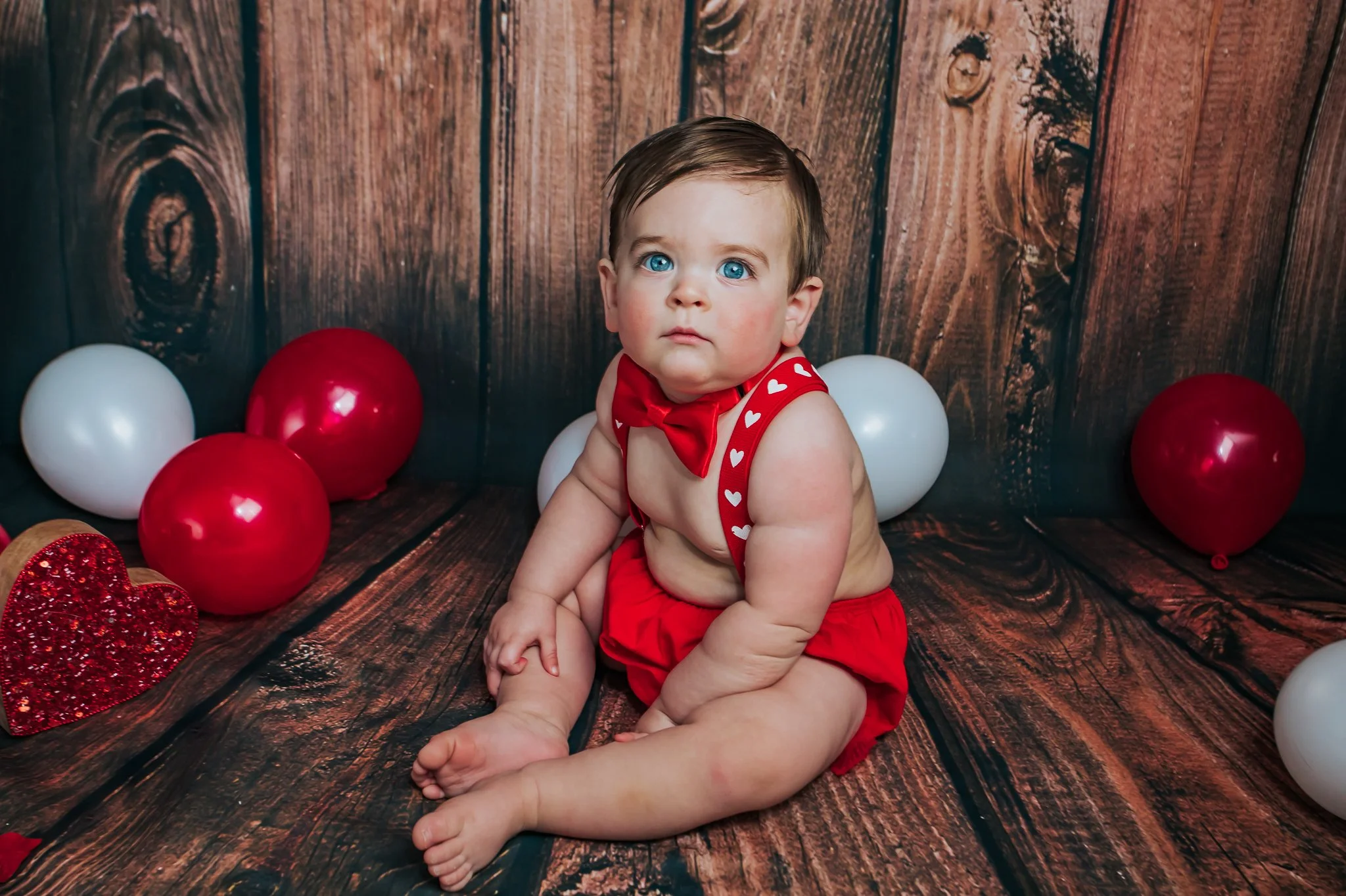 A young child with blue eyes and dark hair, sitting on a wooden floor surrounded by red, white, and glittery hearts and balloons, wearing a red outfit with a heart-patterned red bow tie, looking directly at the camera