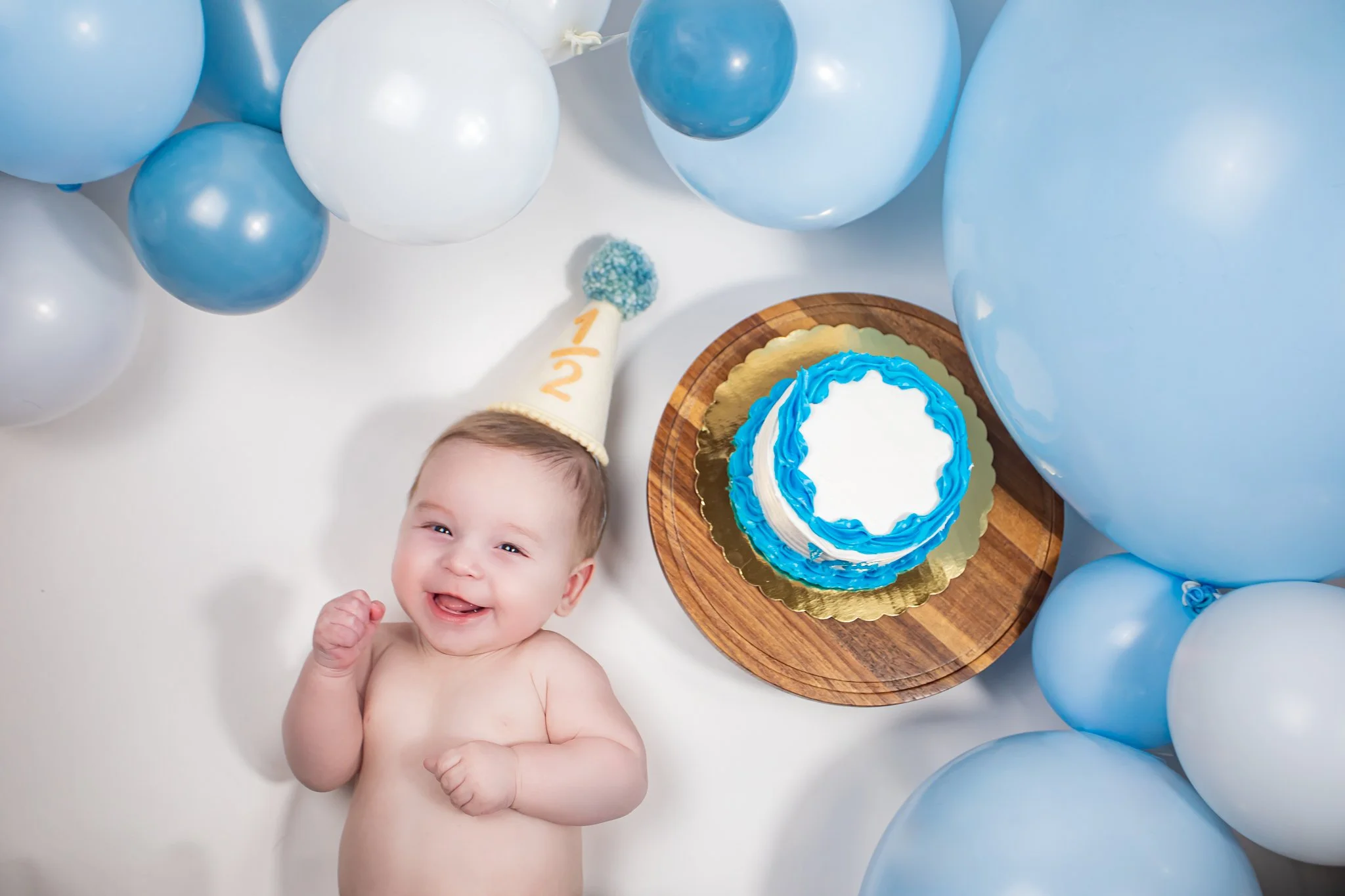 A smiling baby lying among blue and white balloons, a birthday cake with blue and white icing, and a party hat with the number 1/2 on it.