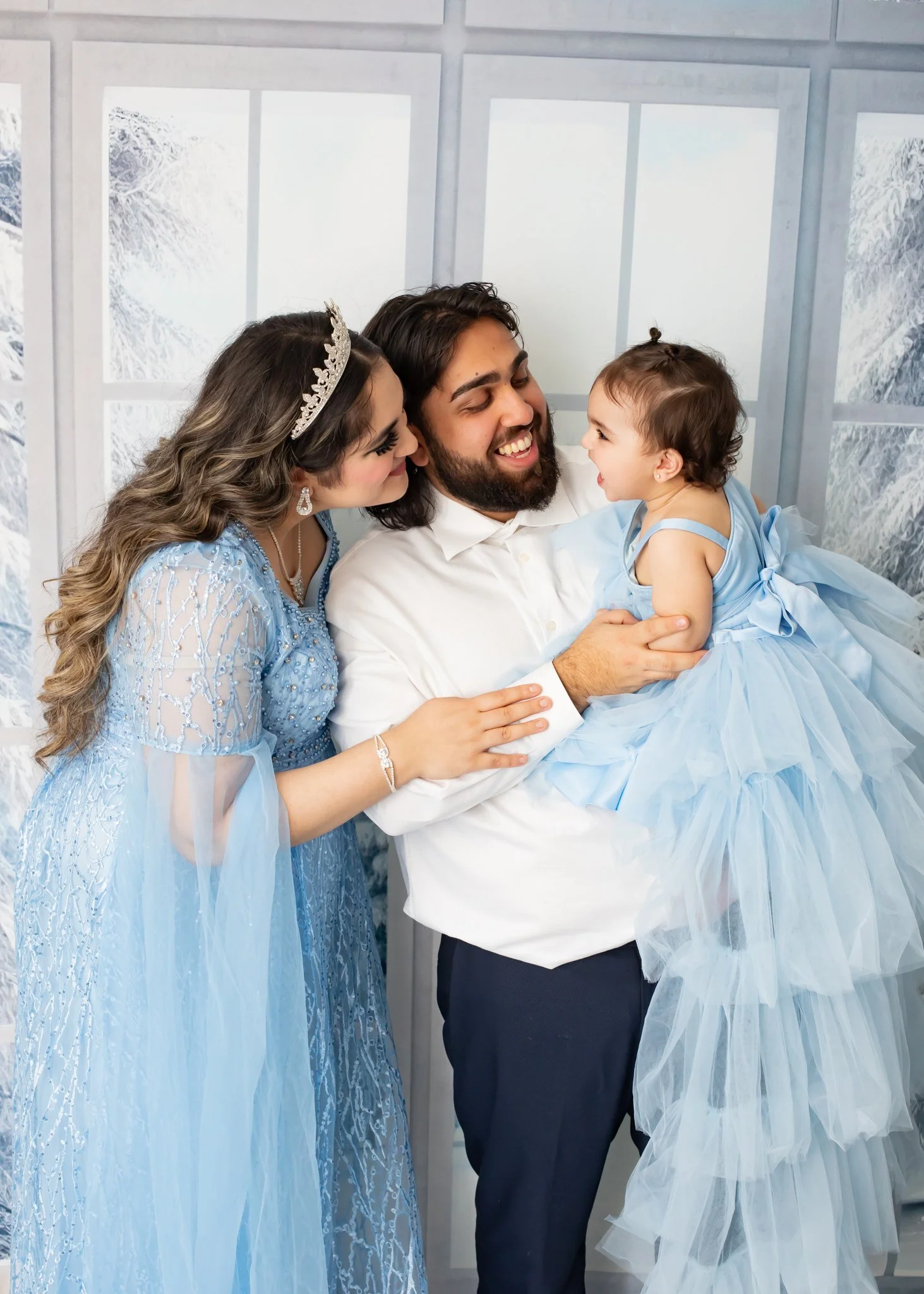 A man, woman, and young girl dressed in blue, celebrating a special occasion, possibly a wedding or christening, in front of a white and silver background.