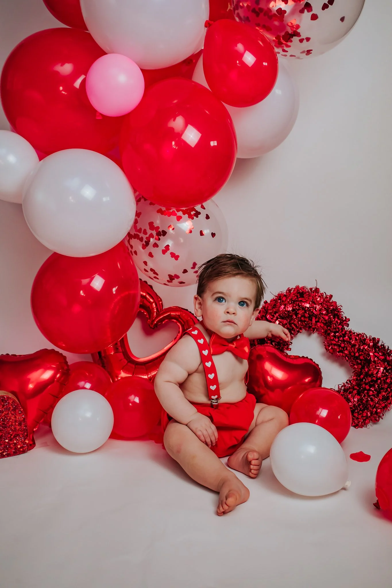 A baby with blue eyes sitting amongst red, white, and clear balloons, some with red heart confetti, in front of a white wall decorated with red balloons and heart-shaped decorations.