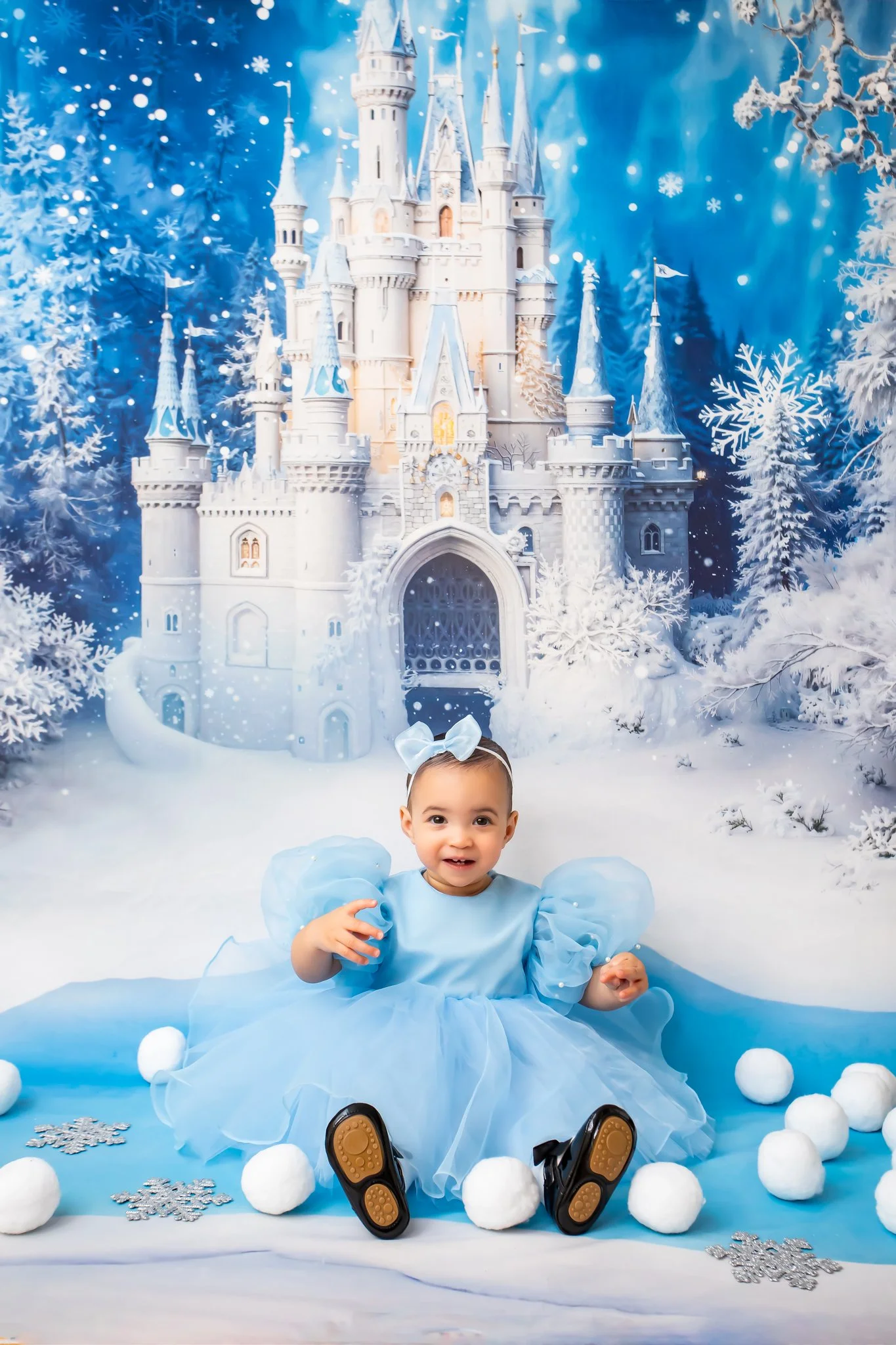 A young girl dressed in a blue princess gown sitting on a snowy white surface surrounded by white snowballs and silver snowflake decorations. In the background, there is a large, illuminated indoor winter scene featuring a castle with blue roofs, sno