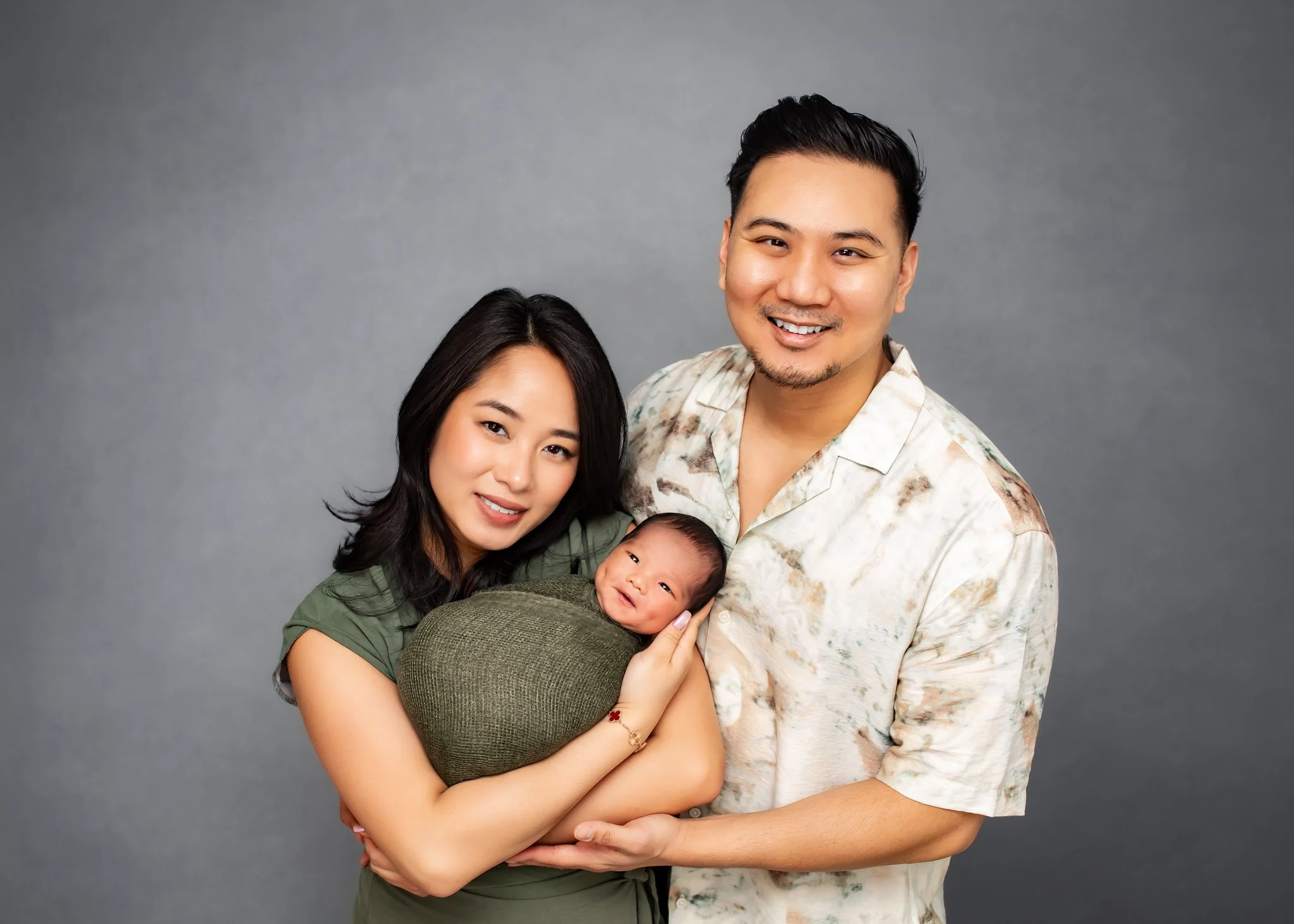 A happy family portrait featuring a woman, a man, and a baby, with the woman holding the baby and all smiling at the camera against a plain gray background.