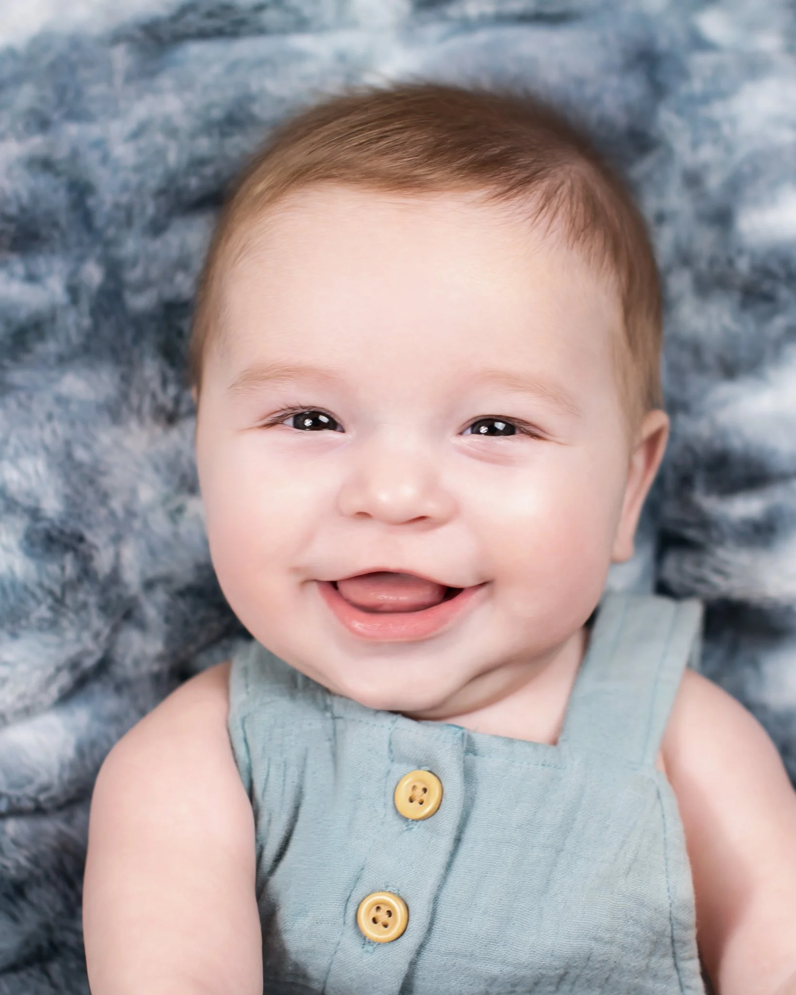 Close-up of a smiling baby with light brown hair, wearing a sleeveless blue shirt with yellow buttons, lying on a gray, fluffy blanket.