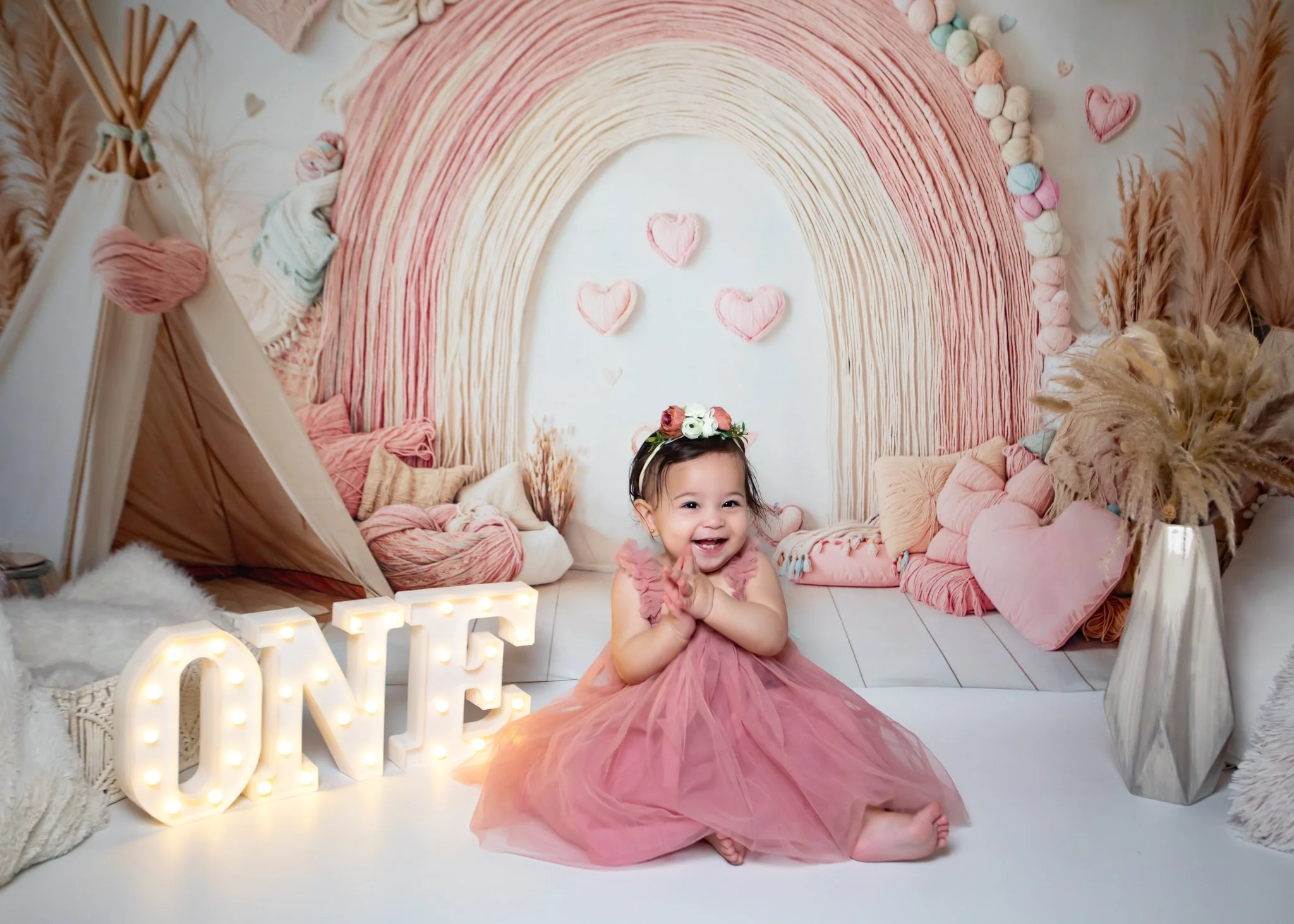 A young girl in a pink dress clapping and smiling in a decorated room with a rainbow backdrop, plush cushions, and heart-shaped decorations.