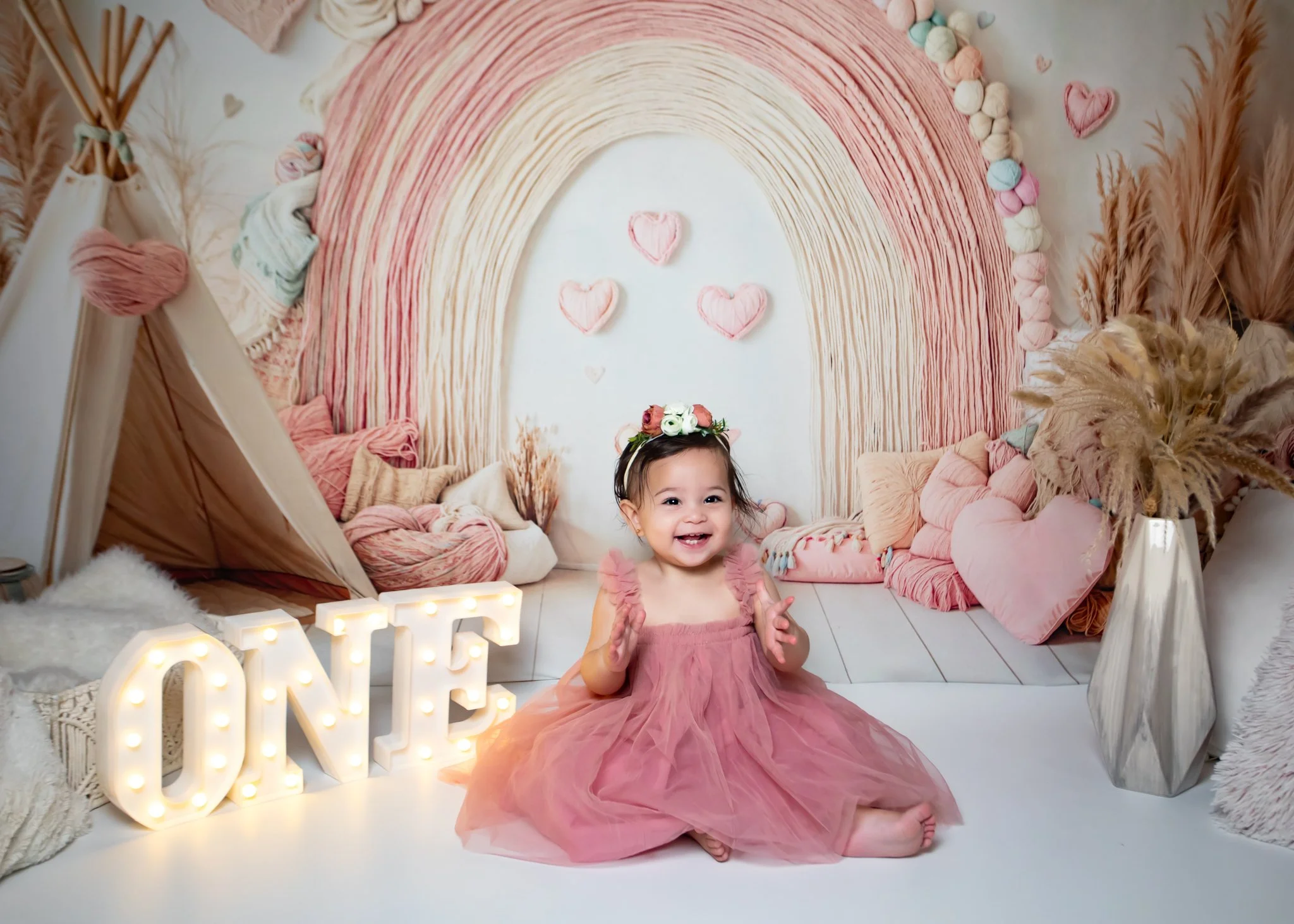 A young girl in a pink tulle dress smiling and clapping in a decorated room with pink, neutral, and earthy tones, a rainbow backdrop with hearts, and a large illuminated 'LOVE' sign.