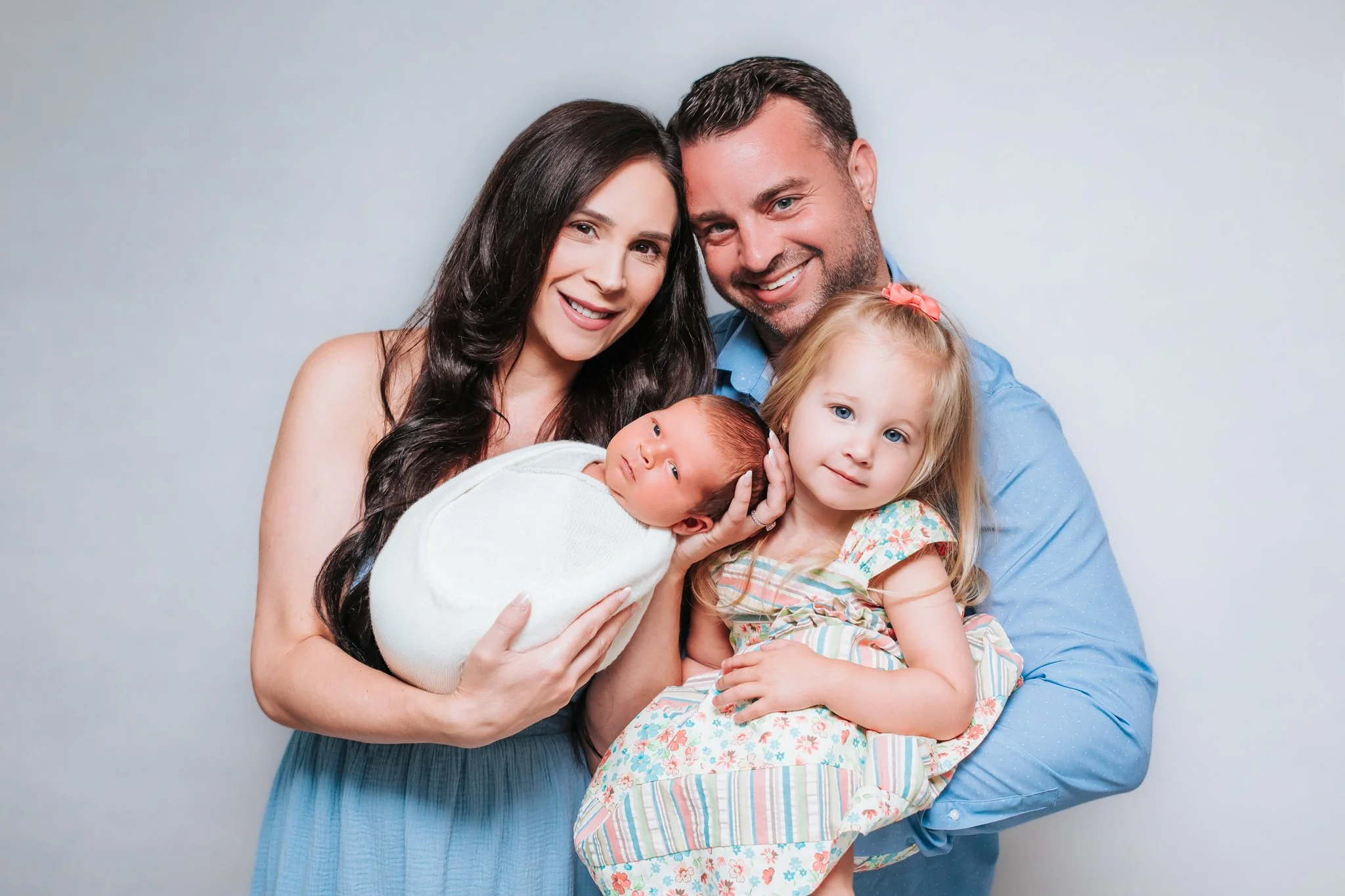 Family portrait of a mother, father, and three children, two girls and a newborn, smiling together against a light gray background.