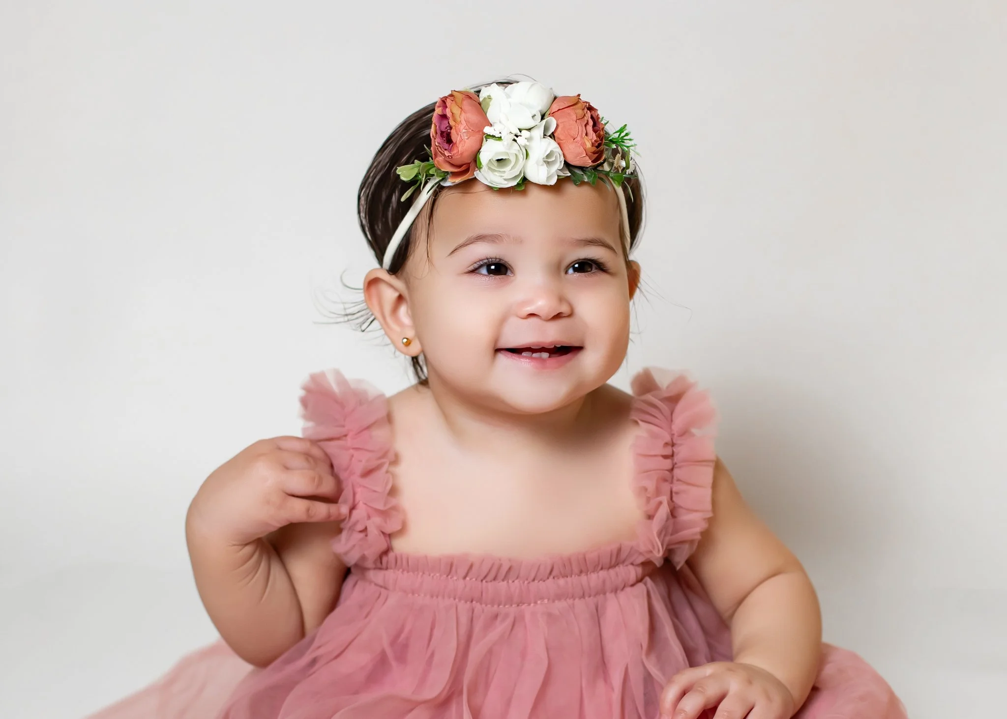 A young girl with dark hair wearing a pink dress and a floral crown with pink, white, and green flowers, smiling and holding her dress with one hand against a plain white background.