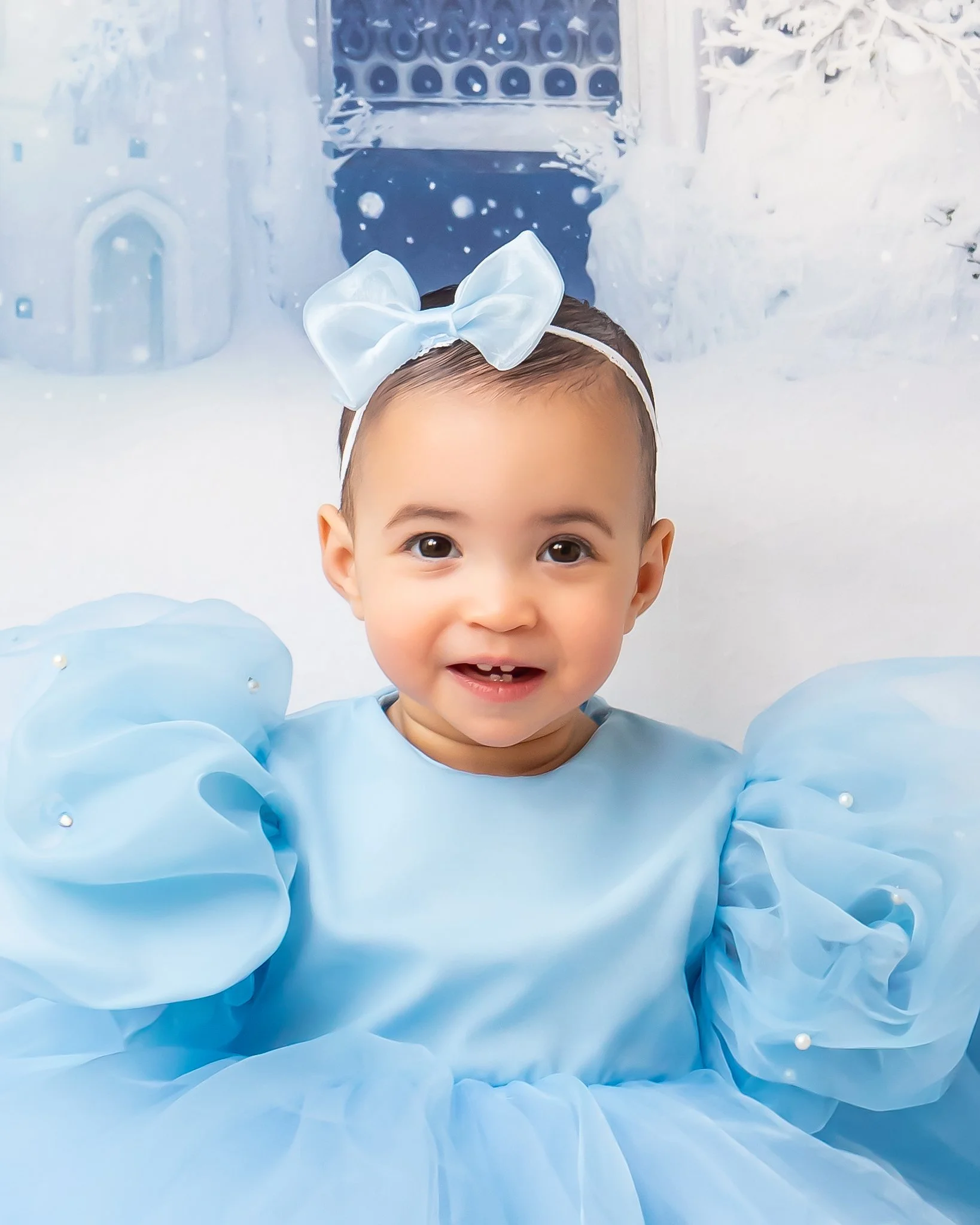 Close-up of a smiling baby girl wearing a large white bow headband and a blue dress with puffy sleeves decorated with small pearls, standing in front of a snowy holiday scene with Christmas decorations and a gingerbread house.