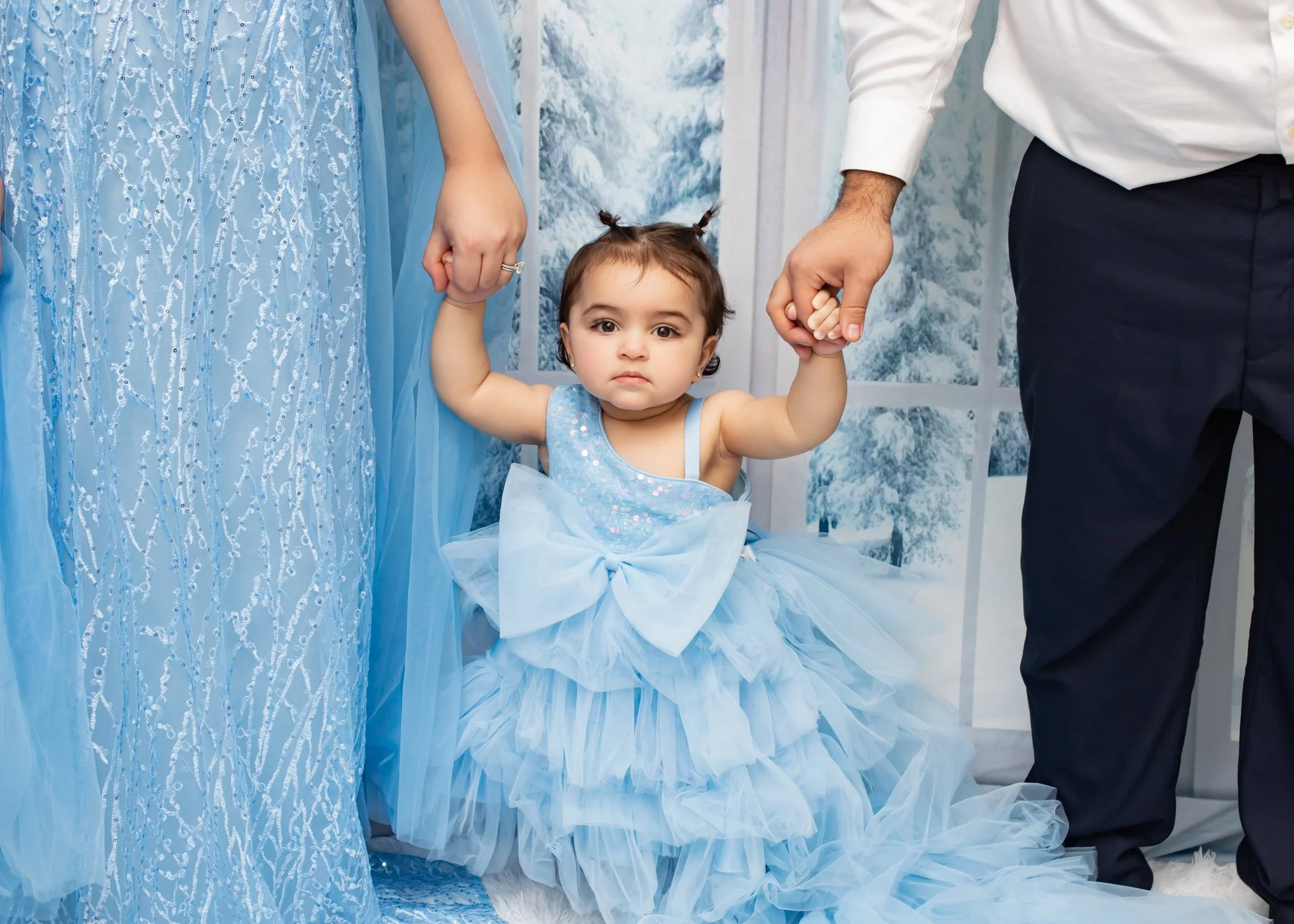A young girl in a blue dress with a tulle skirt and a bow, holding hands with two adults dressed in formal attire, possibly at a special event or celebration in a decorated venue.