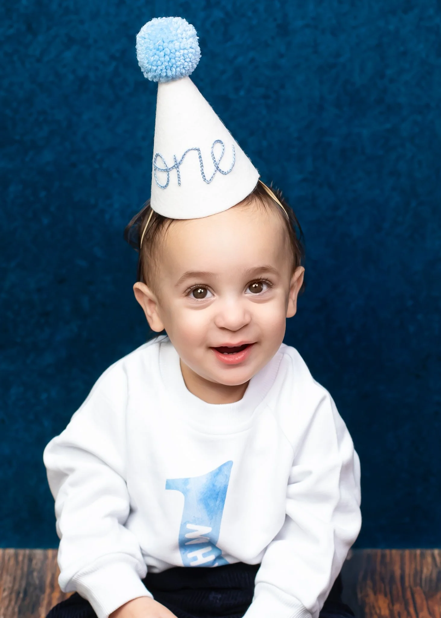 A young boy with brown eyes and brown hair wearing a white sweatshirt with a blue design and a white party hat with a blue pom-pom on top, sitting on a wooden floor with a blue background.