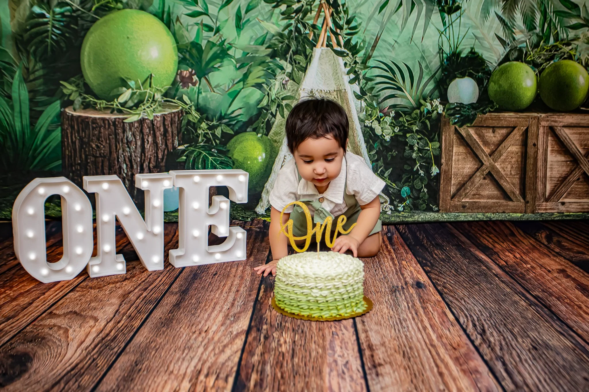 A young boy at a birthday celebration, sitting on a wooden floor, cutting a cake with a yellow 'One' cake topper, with illuminated marquee letters spelling 'ONE' nearby, against a jungle-themed backdrop with large green fruits and foliage.