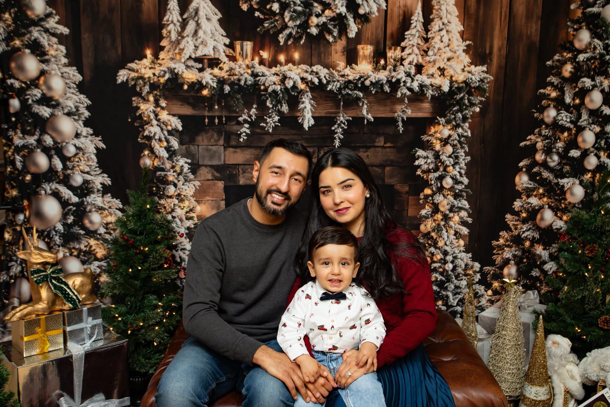 A family of three with a man, woman, and young boy smiling and sitting together in front of a decorated Christmas fireplace with snow-flocked trees, ornaments, and wrapped gifts around them.
