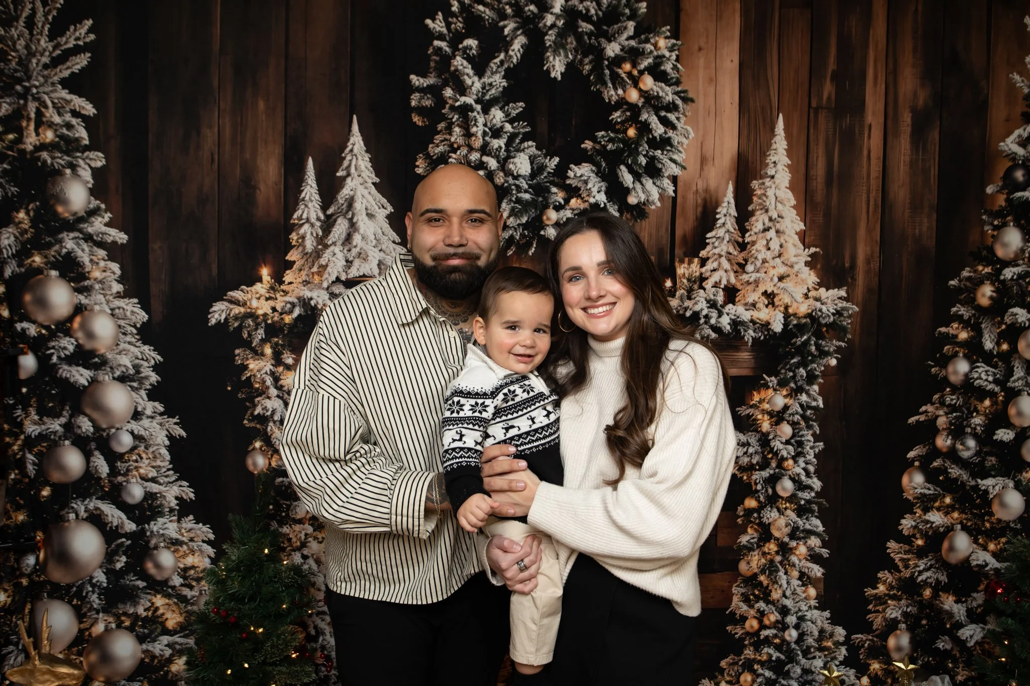 A family of three, including a man, woman, and young boy, smiling and holding each other in front of snowy decorated Christmas trees.