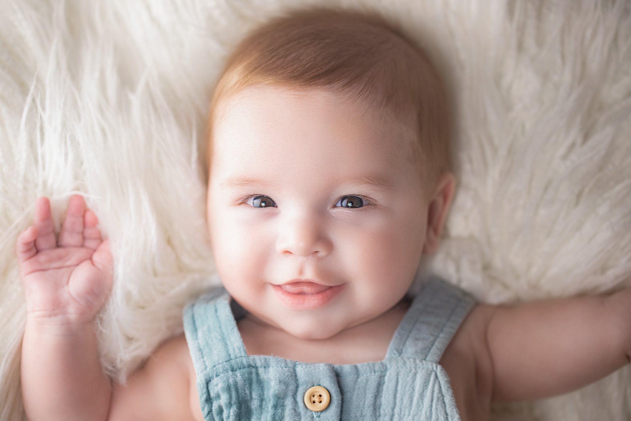 Close-up of a smiling baby with blue eyes, light skin, and light brown hair, lying on a soft, fluffy light-colored surface.
