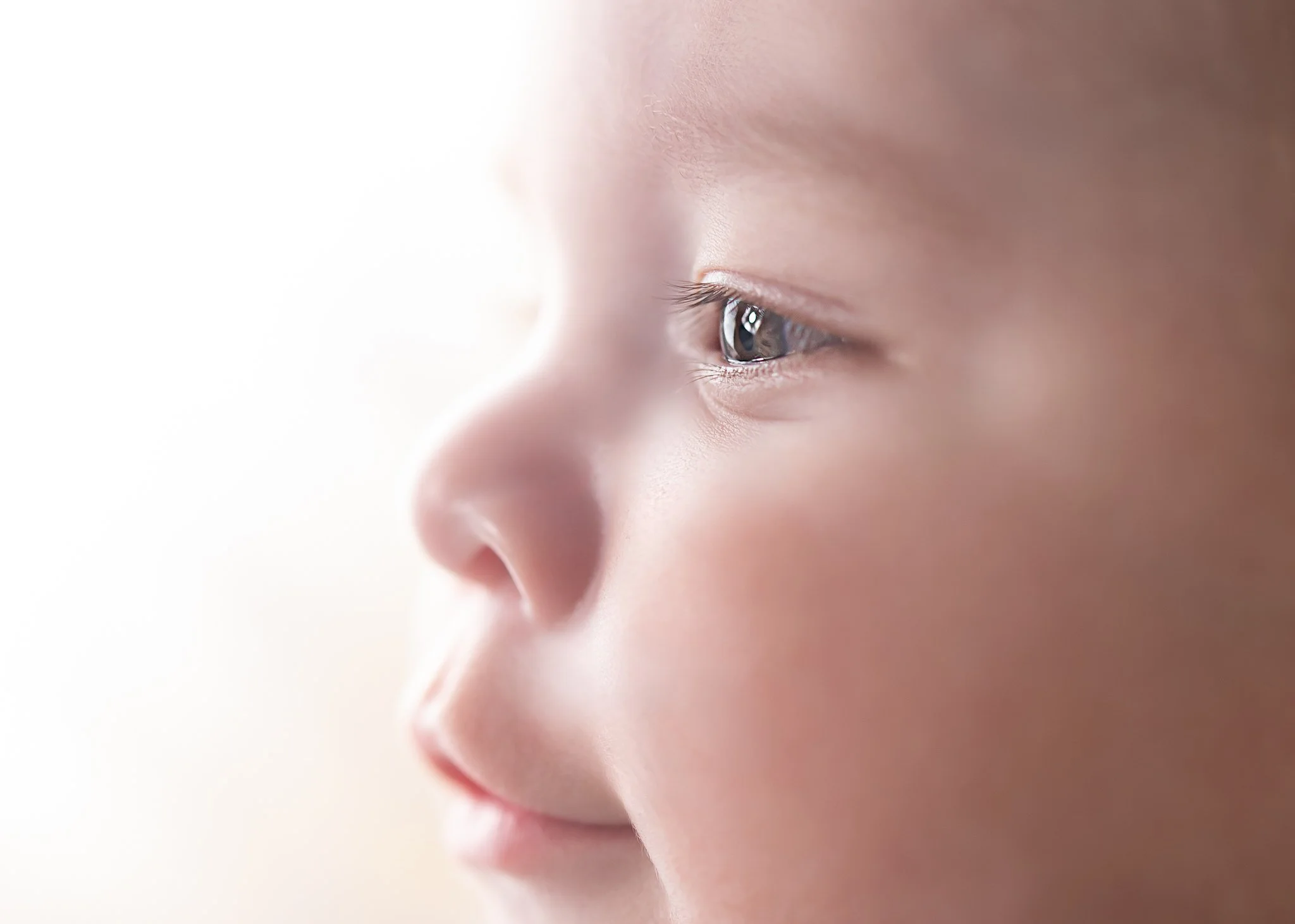 Close-up side profile of a baby's face showing eye, nose, and cheek, with soft lighting and blurred background.