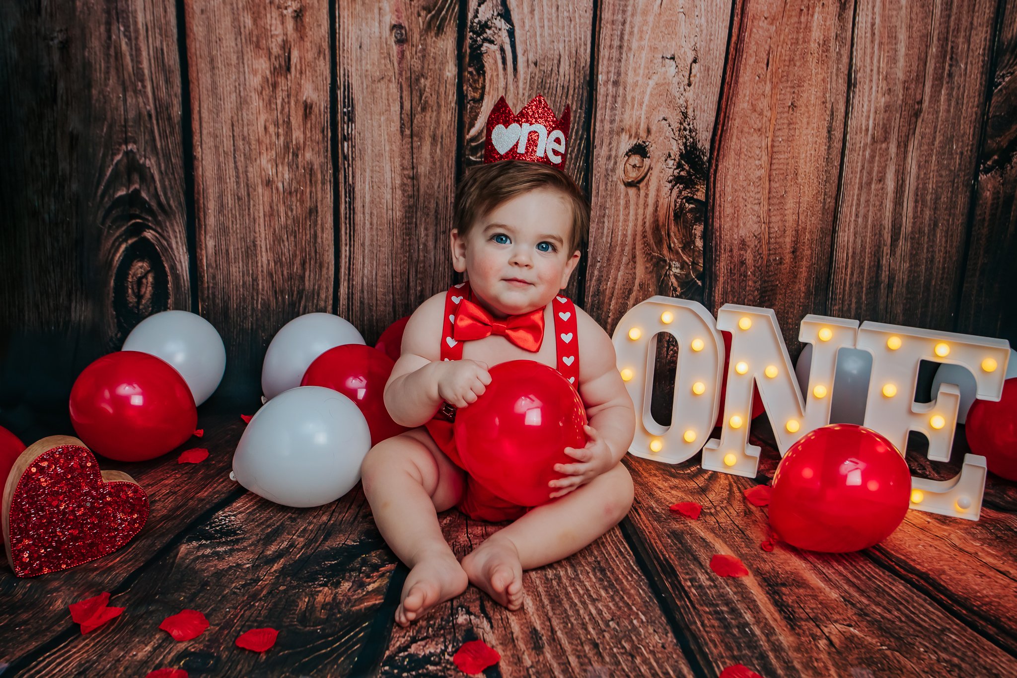 A baby with blue eyes sitting among red, white, and glittery balloons, wearing a red outfit with a heart pattern and a red bow tie, in front of a wooden background decorated with a red glitter crown headband that says "one," illuminated marquee lette