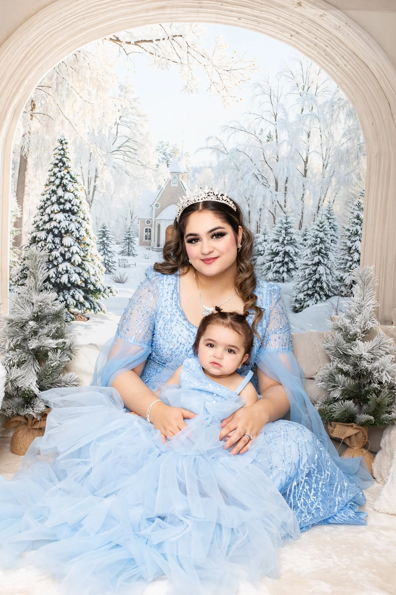 A woman and a young girl dressed in blue sitting on a beige upholstered couch with a winter scene backdrop of snow-covered trees and a church, with snowflake trees and curtains.