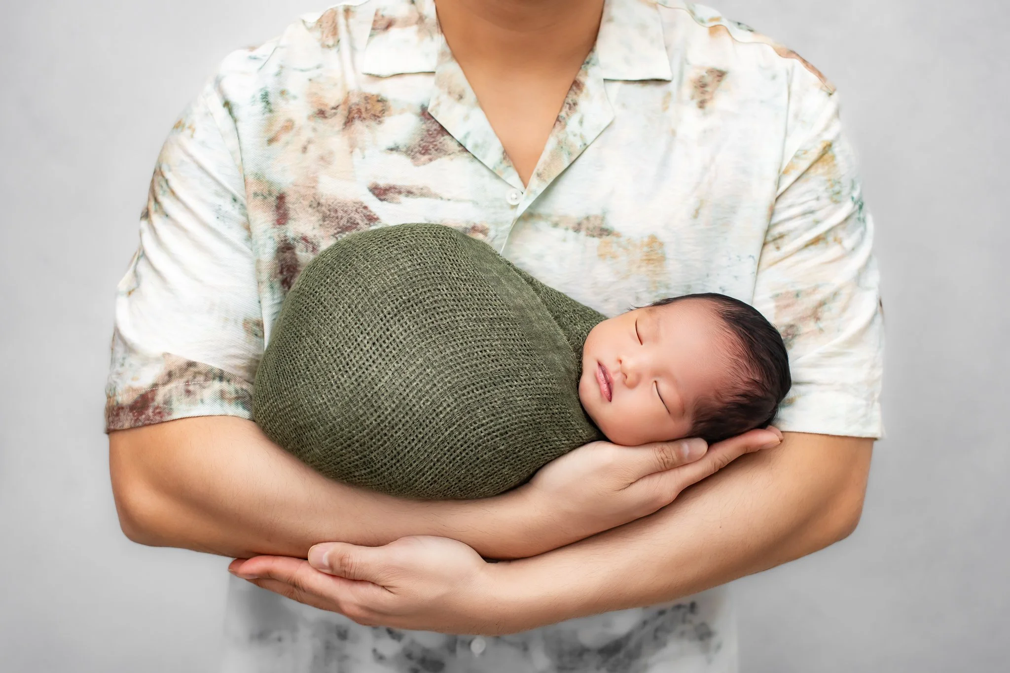 Person holding a sleeping baby wrapped in a green blanket, wearing a floral shirt, against a plain background.