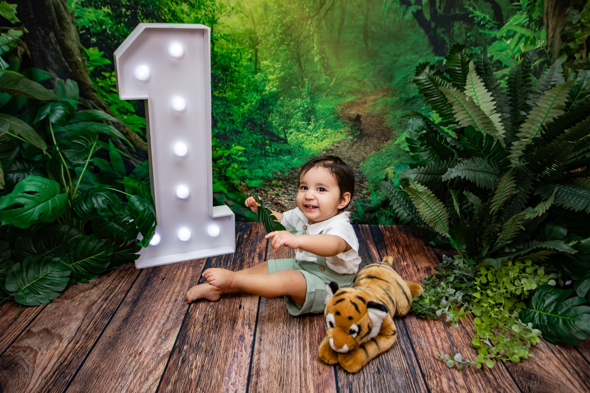 A smiling toddler sitting on a wooden floor surrounded by green lush plants and a forest background, holding a green leaf with a plush tiger toy beside him.